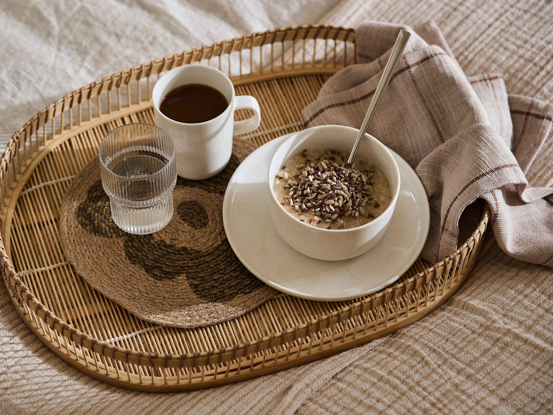 Breakfast in bed with oatmeal topped with seeds, coffee, and water on a rattan tray with a striped napkin.