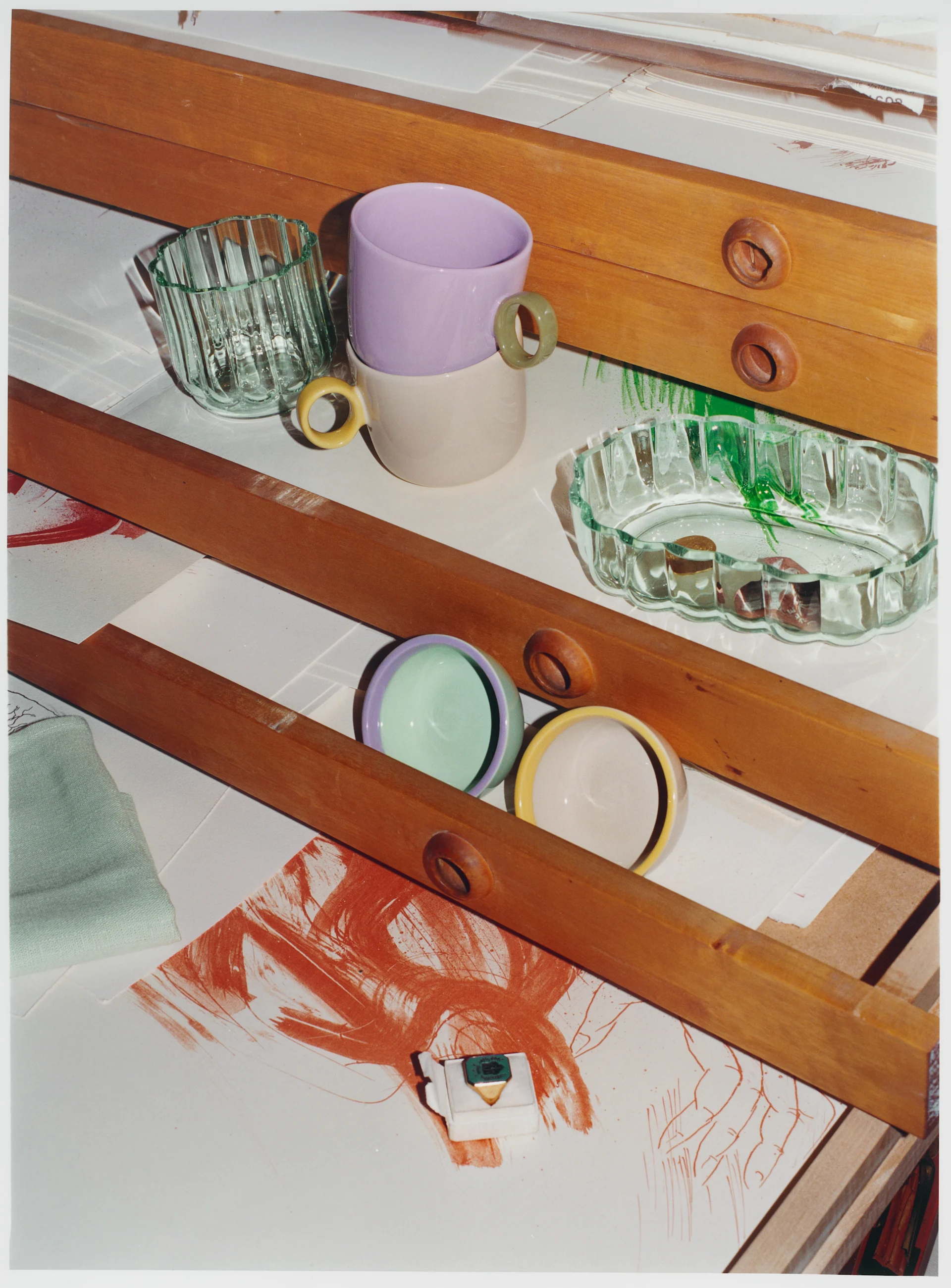 Pastel ceramic cups, clear fluted glassware, and a gold ring with a green stone on a white surface with red brushstrokes and wooden beams.