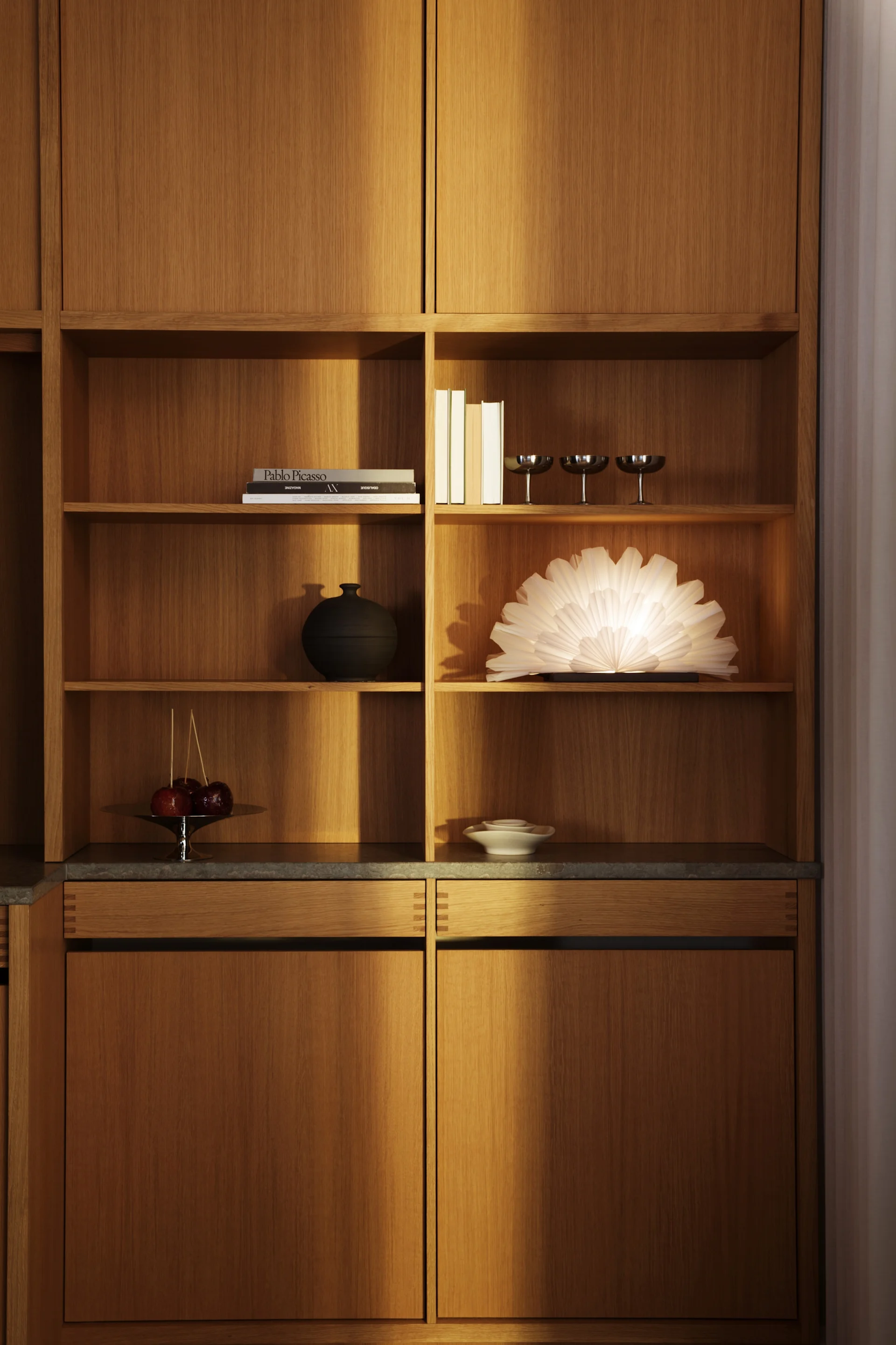 A built-in wooden shelving unit with various decor items including books, silver glasses, a black vase, a glowing white fan lamp, candy apples, and white bowls.