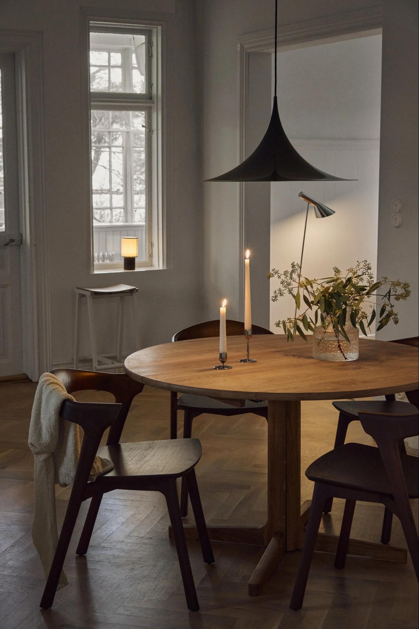 A cozy dining room with a round wooden table, lit candles, a vase with greenery, dark chairs, and a bell-shaped pendant lamp.