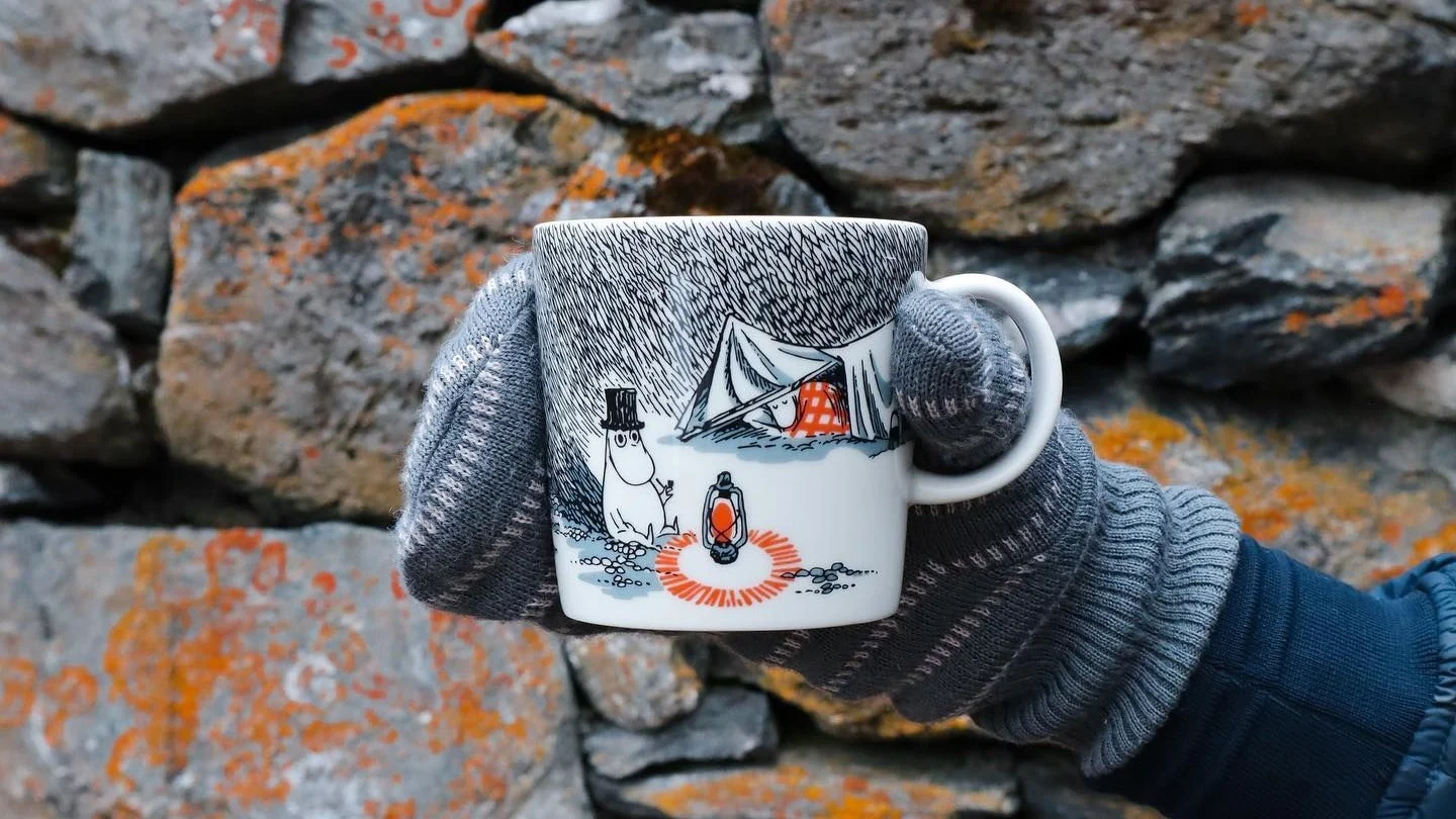 @moominbyingri holds the Schlaf gut Moomin cup in front of a stone wall wearing a glove.