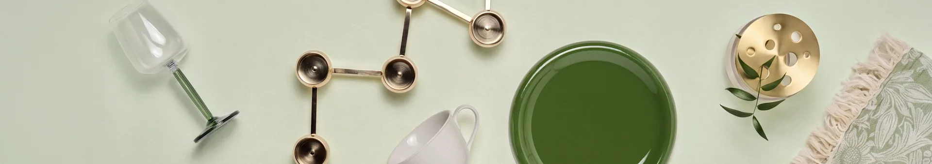 Flat lay: glass with green stem, gold sculptural decor, white mug, green plate, brass item with holes and leaves, and a fringed green patterned textile.