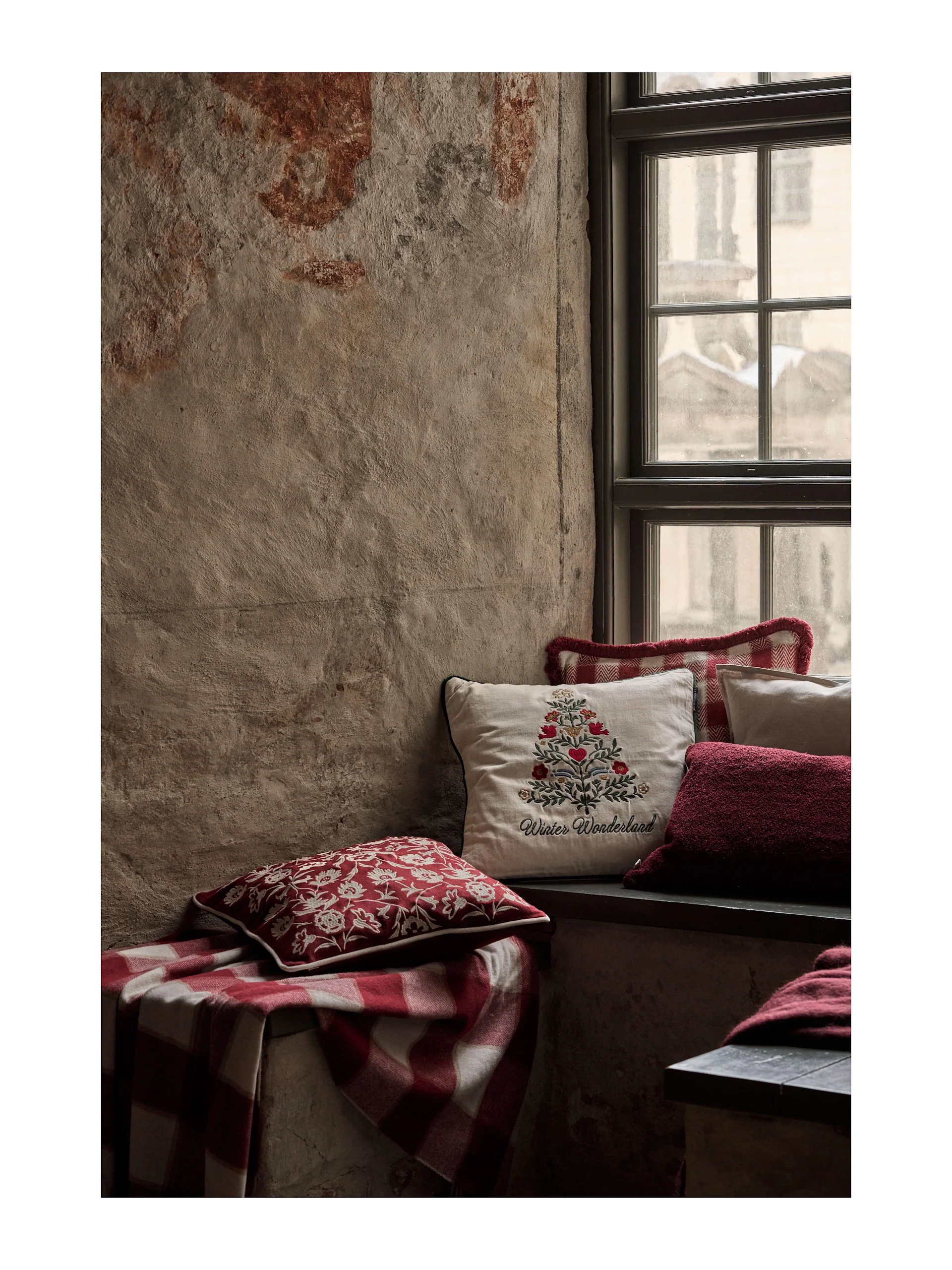 Cozy window seat with festive red and white pillows and a plaid blanket. A cream pillow features 'Winter Wonderland' embroidery.