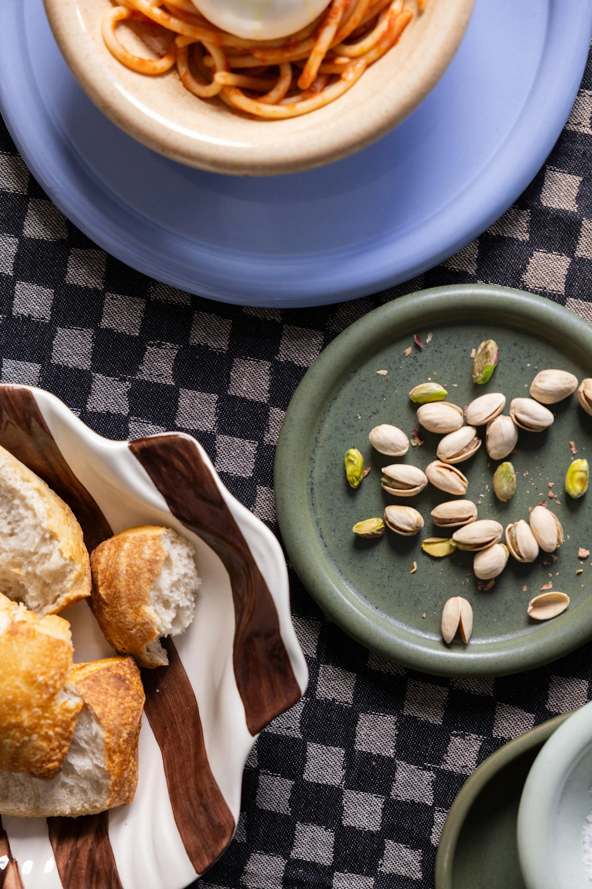 Spaghetti on a blue plate, pistachios on a green plate, and bread in a striped bowl on a checkered tablecloth.
