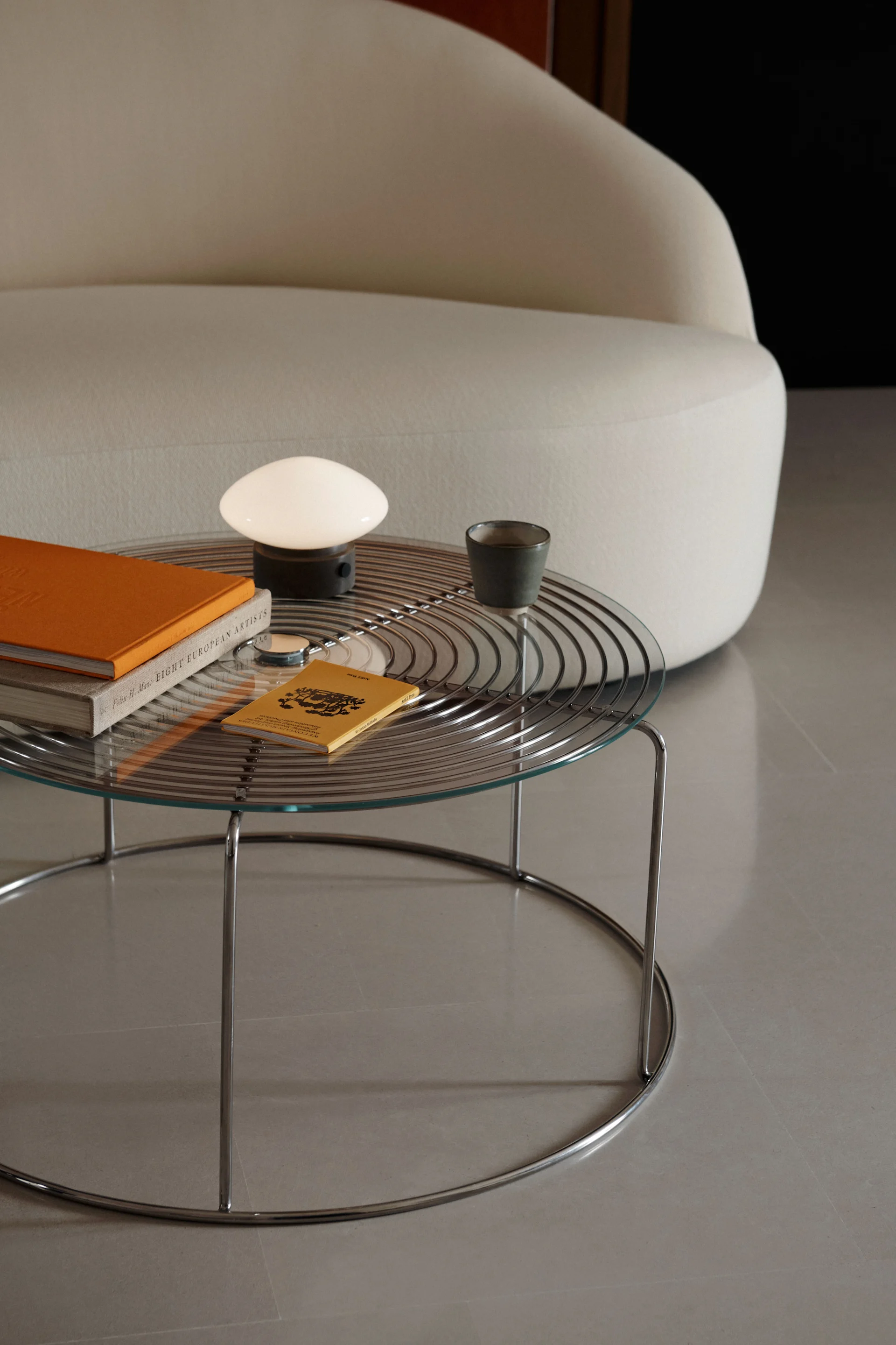 A glass-top coffee table with a chrome base holds a white mushroom lamp, an orange book, and a dark mug next to a curved cream sofa.