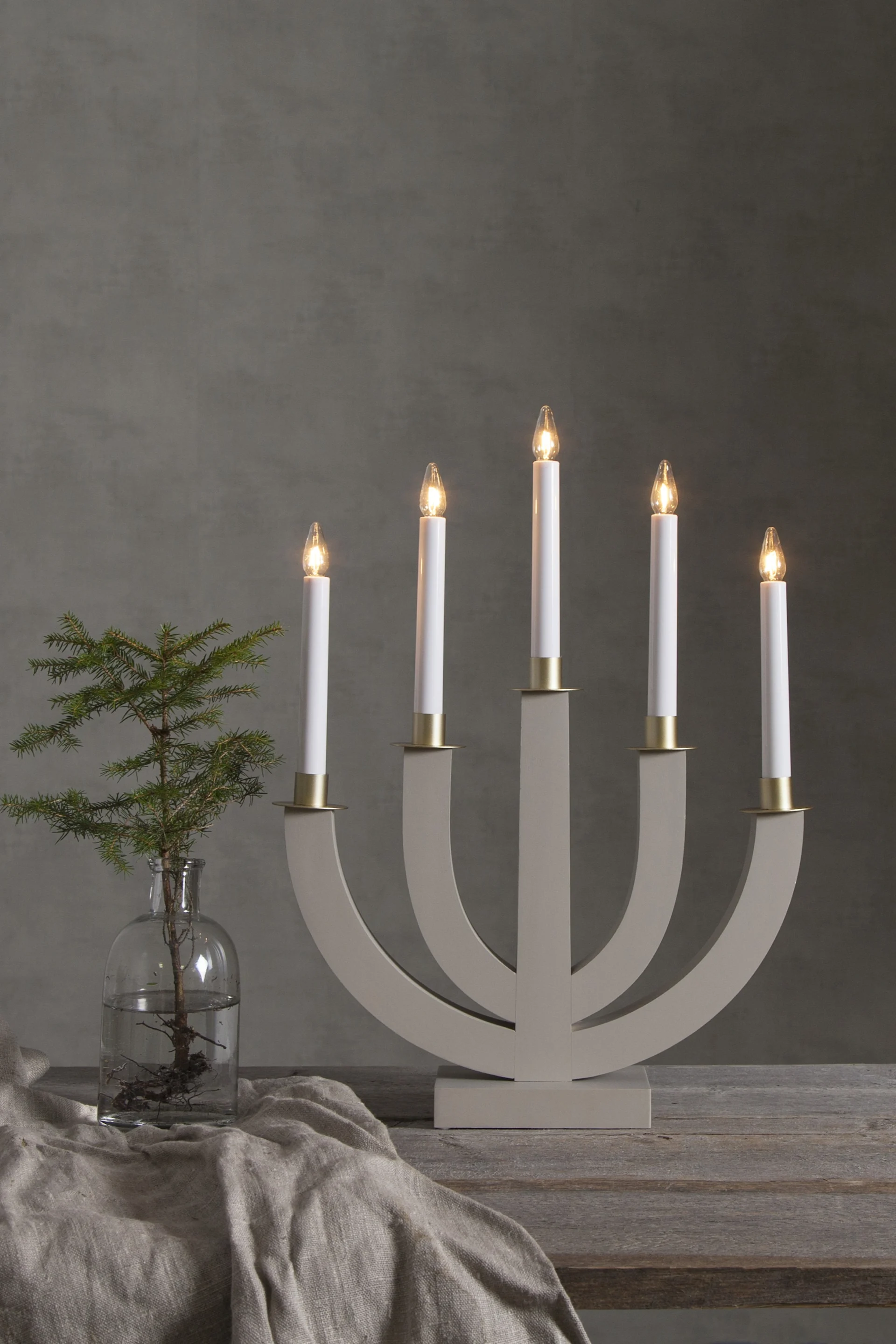 The Eli candle arch from Star Trading in grey stands on a sideboard next to a small vase with a small green branch. 