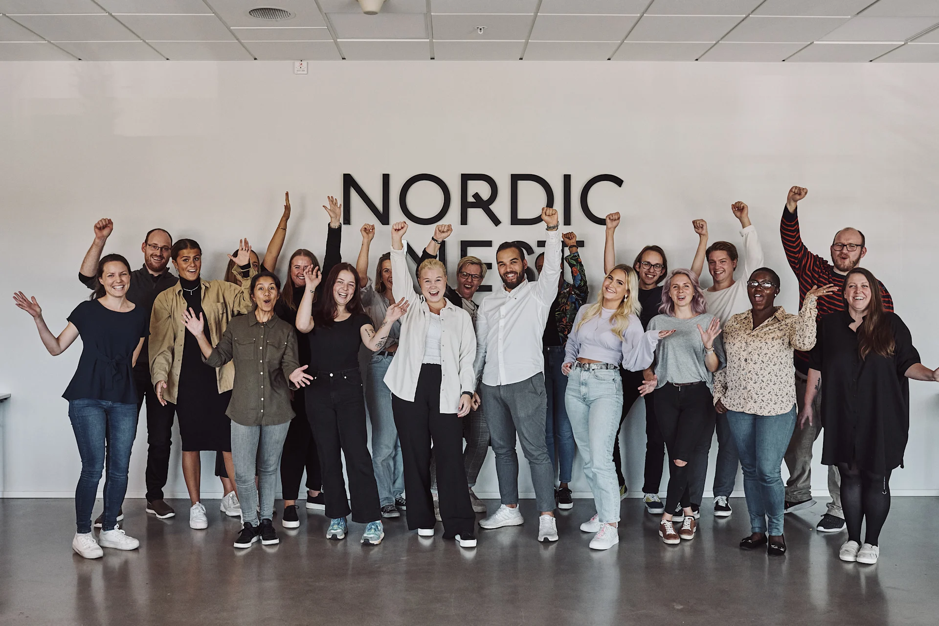A diverse group of smiling people raising their fists in front of a 'NORDIC' sign on a white wall.