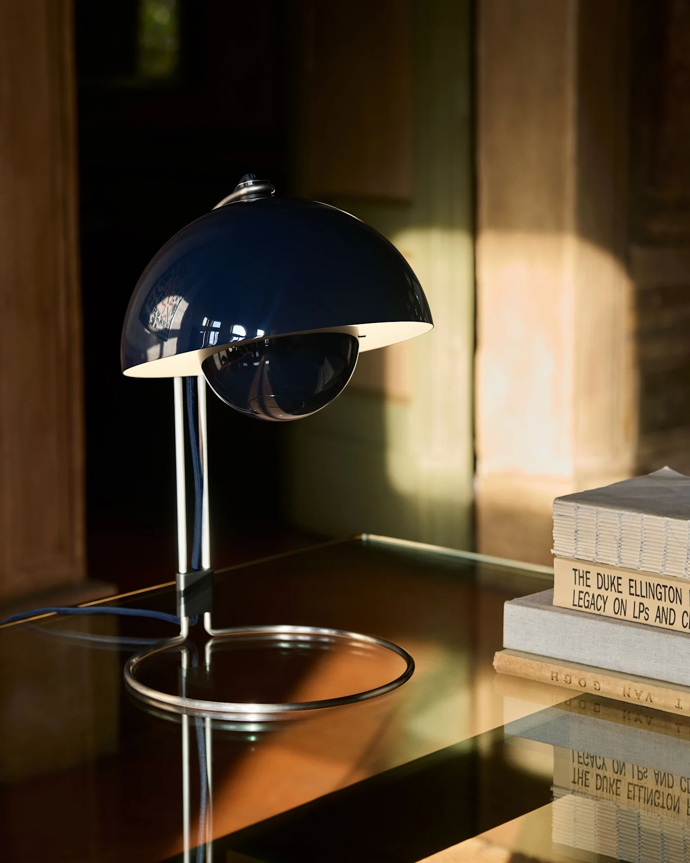 A dark blue, bell-shaped table lamp with a chrome base sits on a reflective table next to a stack of books.