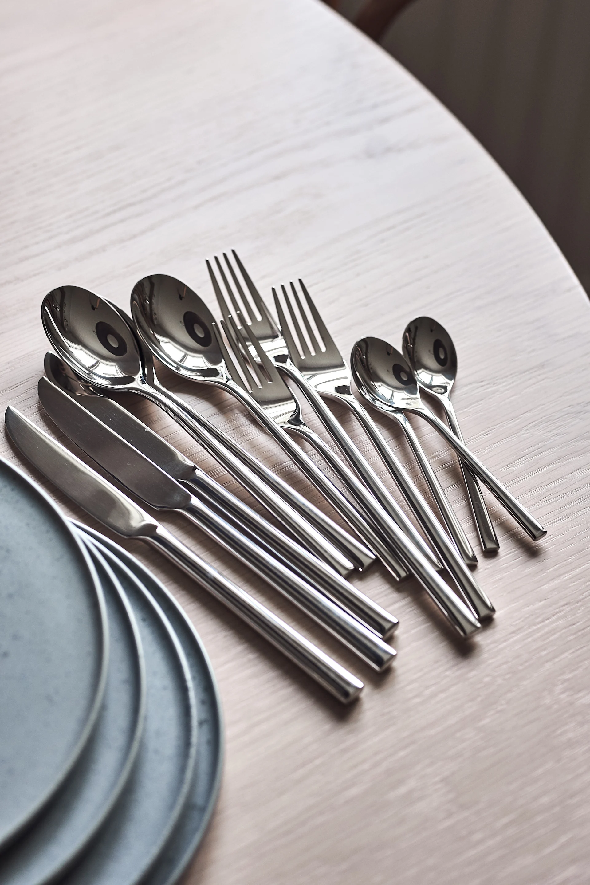 Set of shiny silver-colored cutlery including spoons, forks, and knives, arranged on a light wooden table with grey plates.