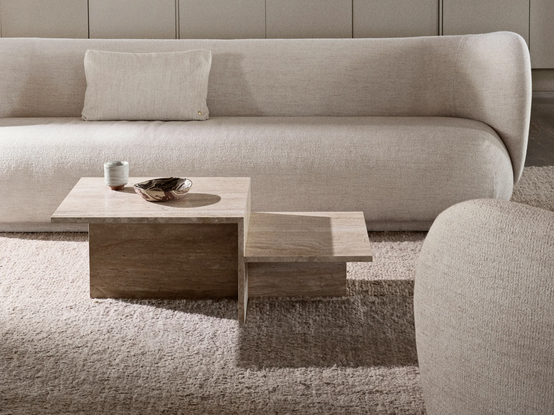A cream textured sofa with a matching pillow, two stacked travertine coffee tables, and a shaggy cream rug.