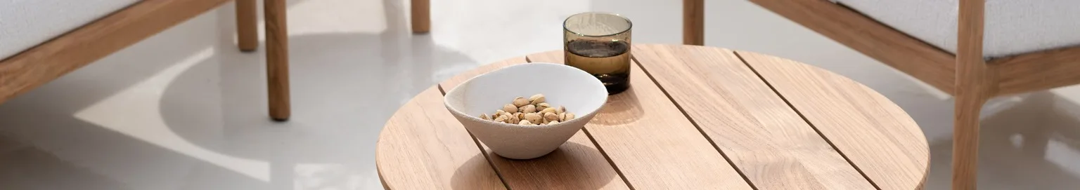 A round wooden side table with a white bowl of pistachios and a glass of water, next to wooden chairs with white cushions.