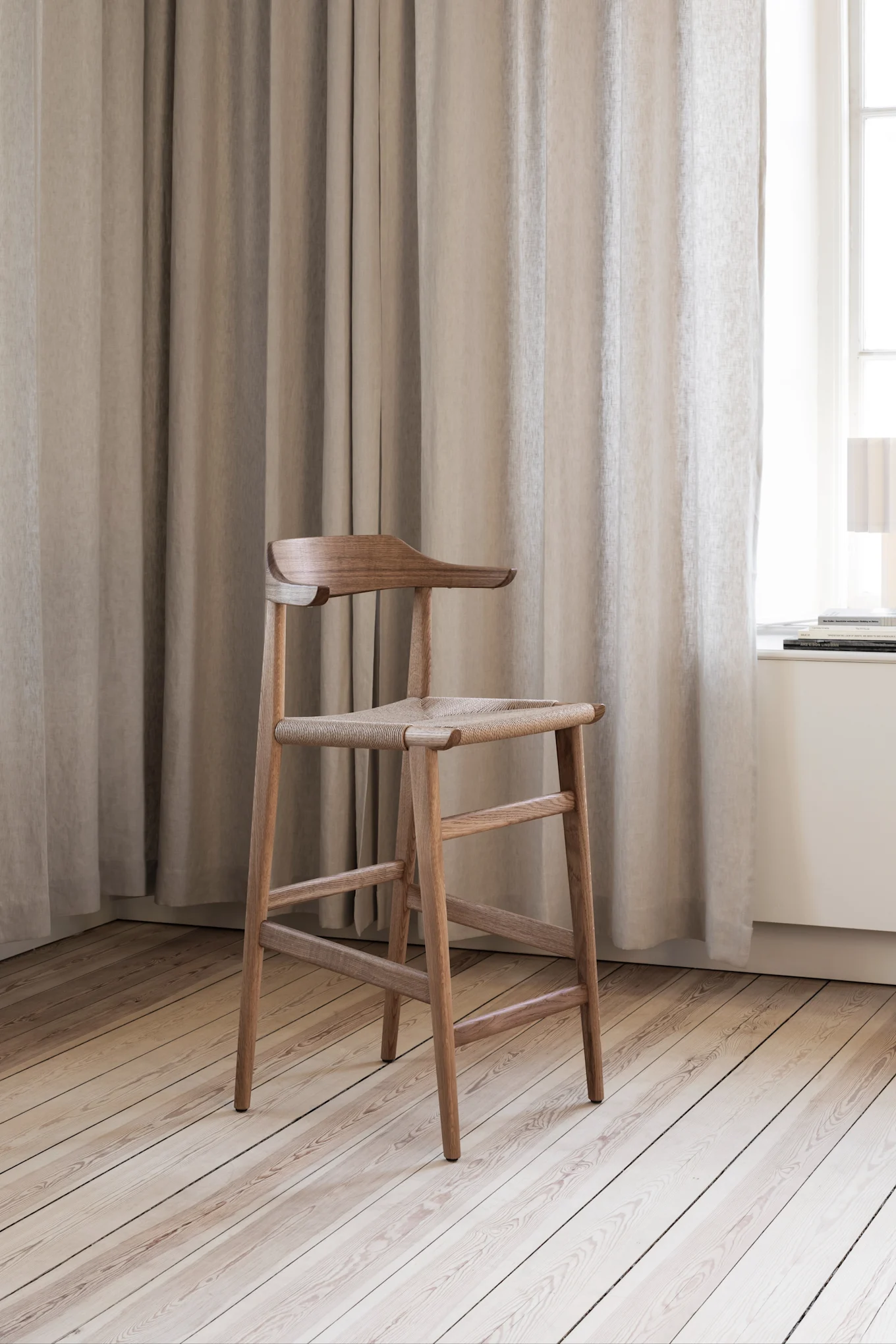 A wooden counter-height stool with a woven seat and curved backrest, in a room with light curtains and a wooden floor.