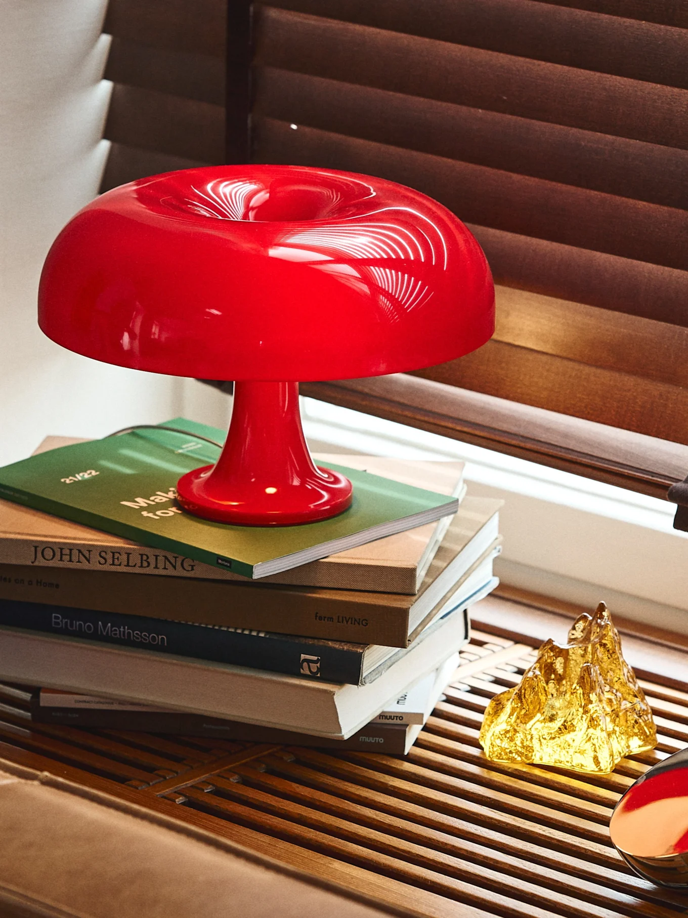 A glossy red mushroom-shaped lamp on a stack of books, alongside a golden textured sculpture, with wooden window blinds in the background.