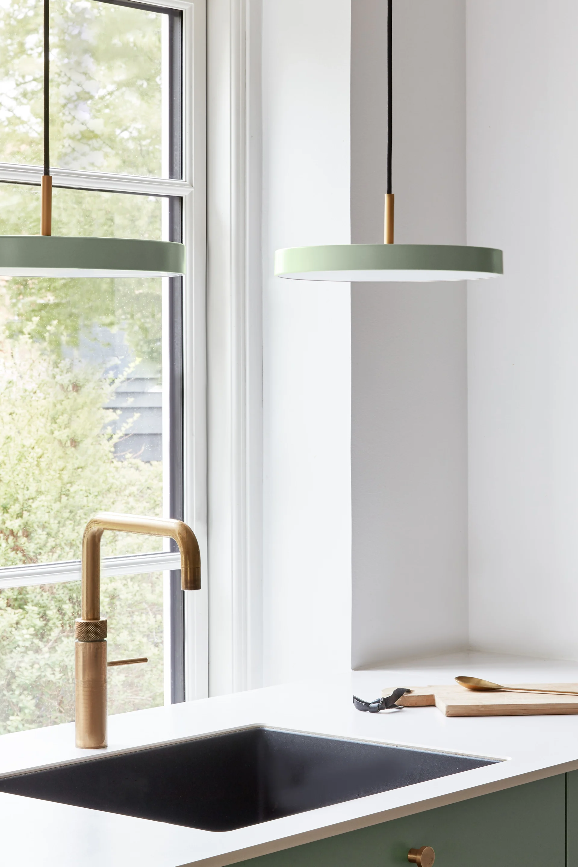 Modern kitchen corner with a black sink, brass faucet, green cabinets, and two pale green pendant lights next to a window.