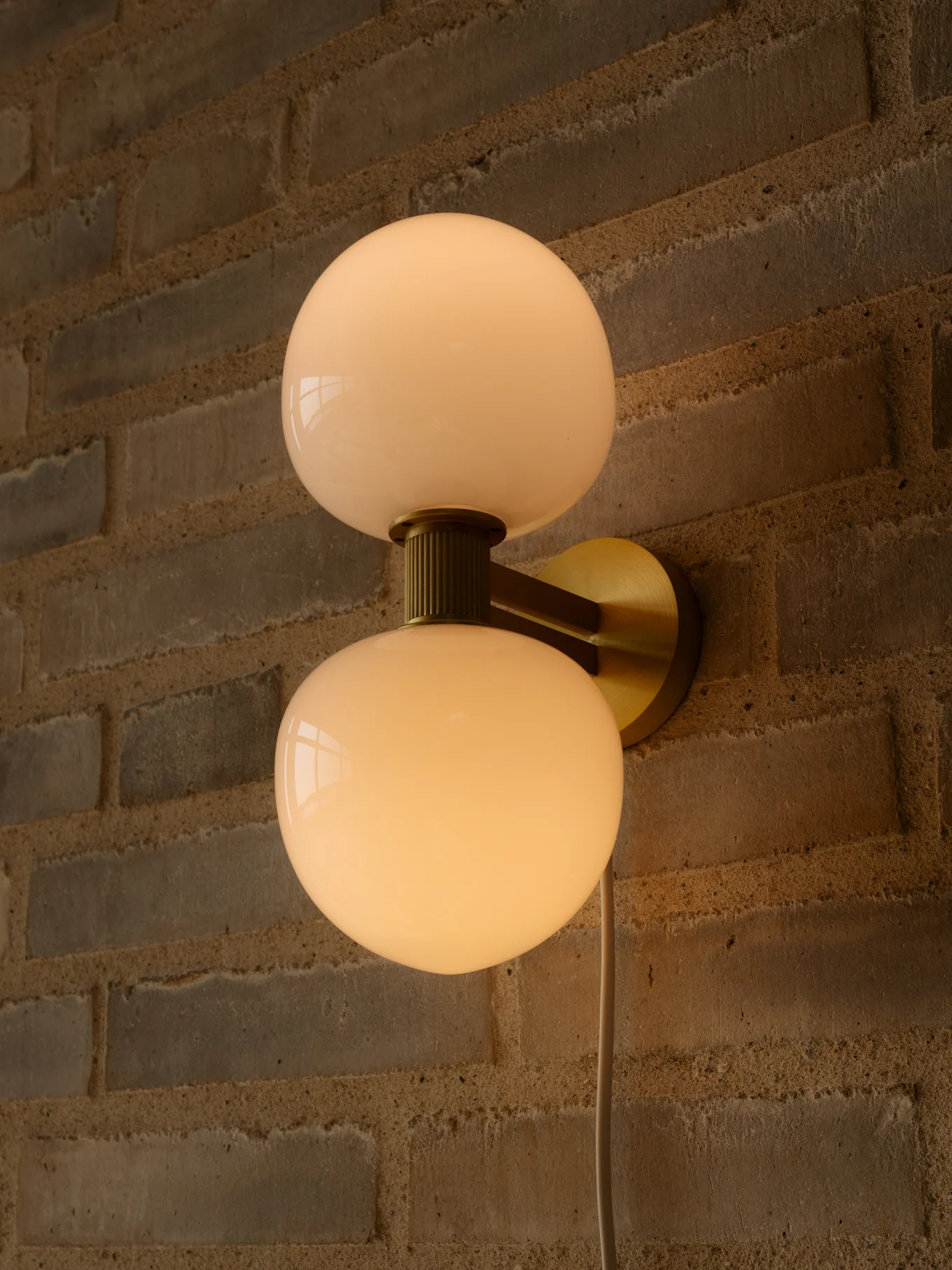 Wall light with two milky white spherical glass shades and a brass metal mount on a brick wall.