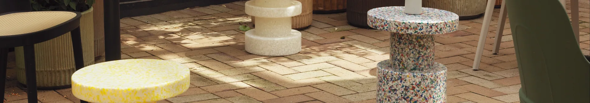 Outdoor patio with a black chair, yellow, beige, and multicolor speckled stools, and a green chair on brick flooring.