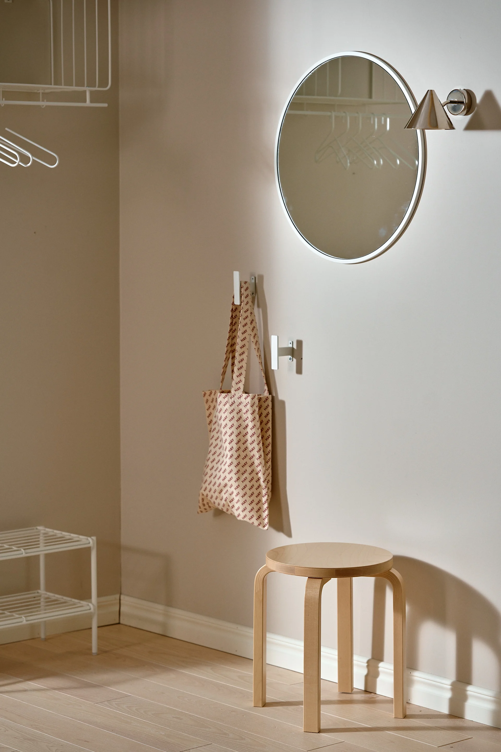 A minimalist entryway with a round mirror, a wooden stool, a 'HAY' patterned tote bag on a hook, and white wire shelves.