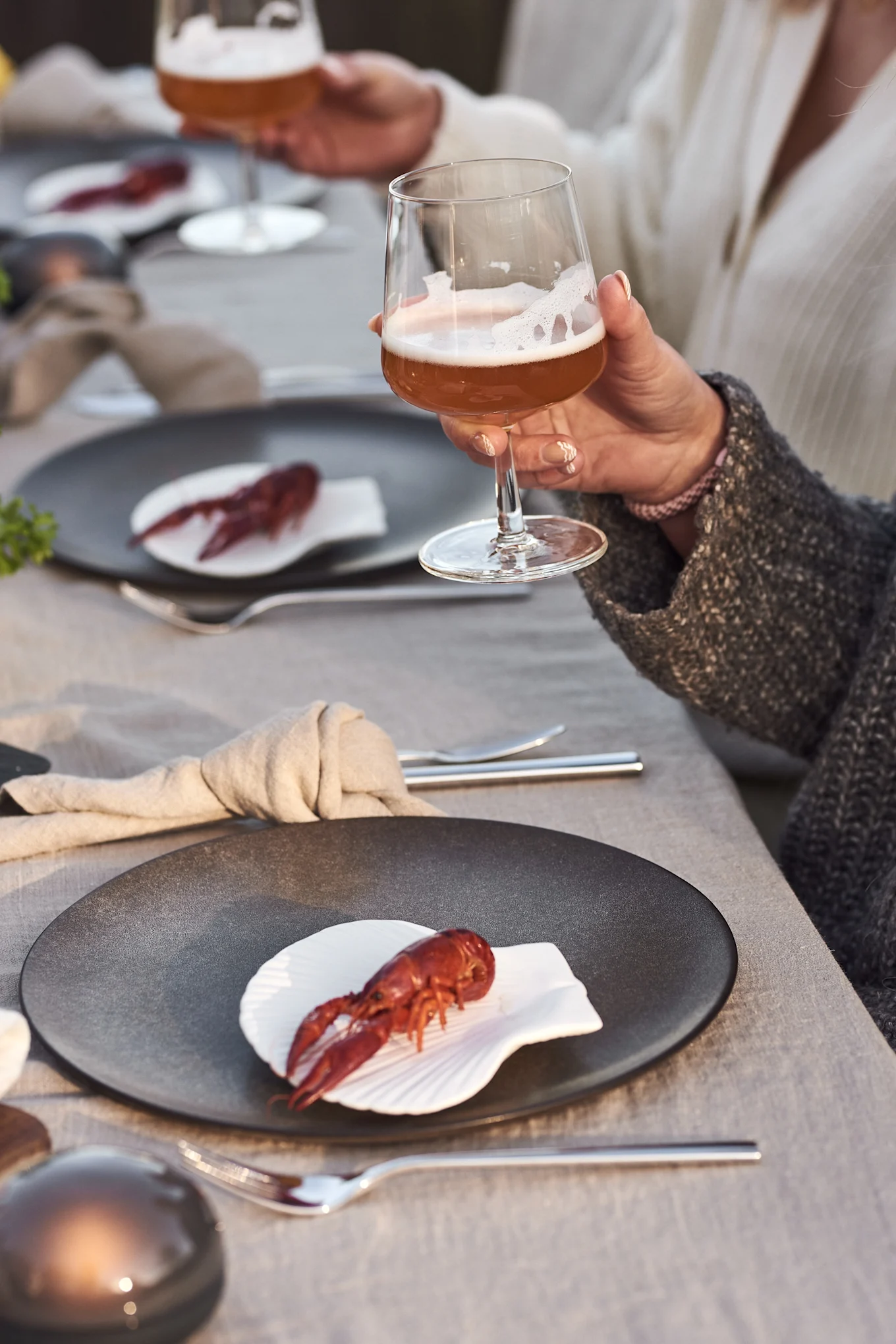A person holds a glass of beer, with a plate of cooked crawfish and silverware on a linen tablecloth.