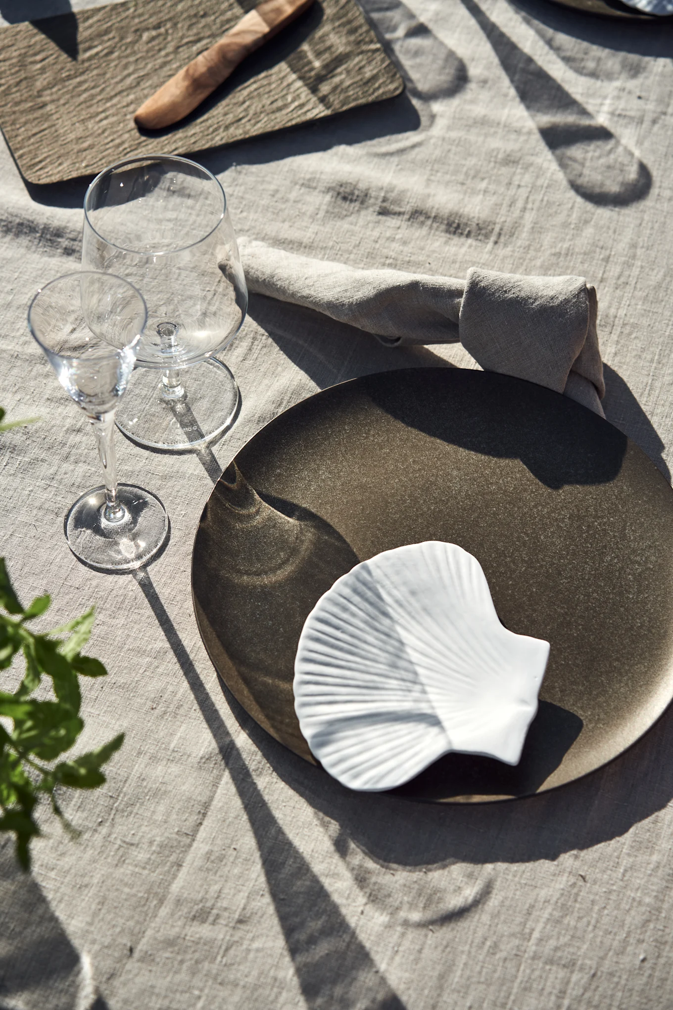 An outdoor table setting with a dark bronze dinner plate, a white shell dish, and two wine glasses on a linen tablecloth, with strong shadows.