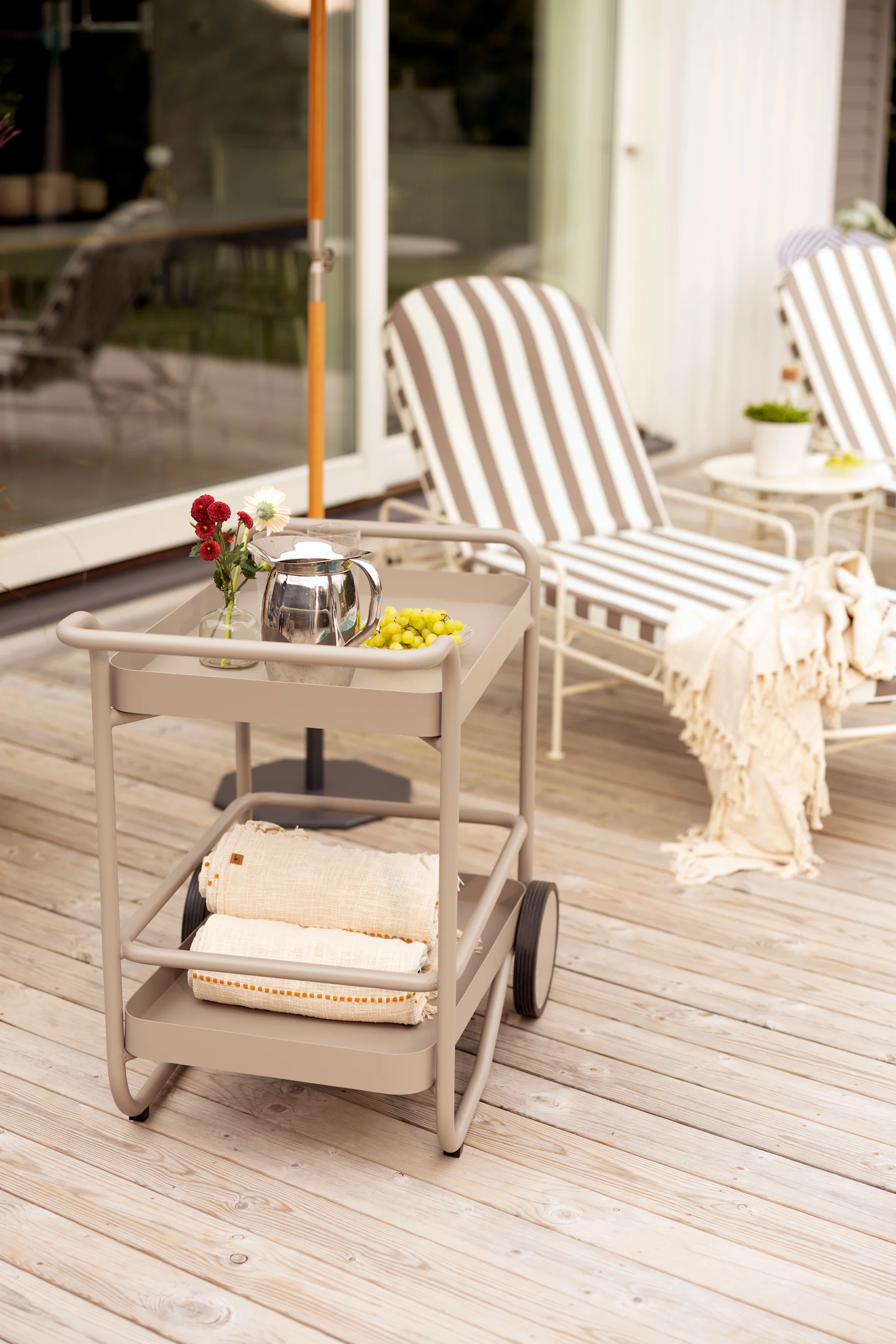 Two-tiered beige outdoor serving cart with a teapot, grapes, and flowers on the top, and rolled towels on the bottom.