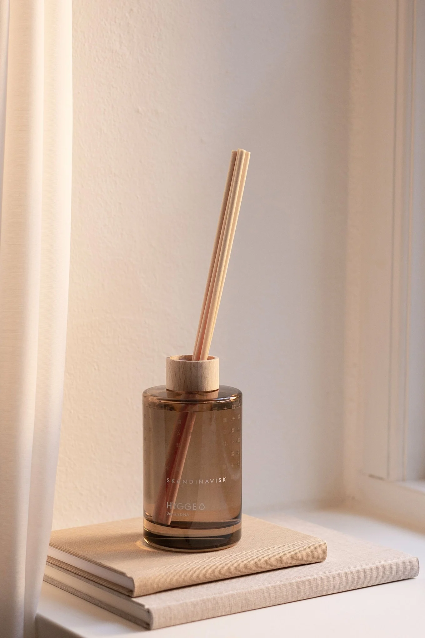 A brown glass reed diffuser with wooden reeds and cap, sitting on two light-colored books on a windowsill next to a curtain.
