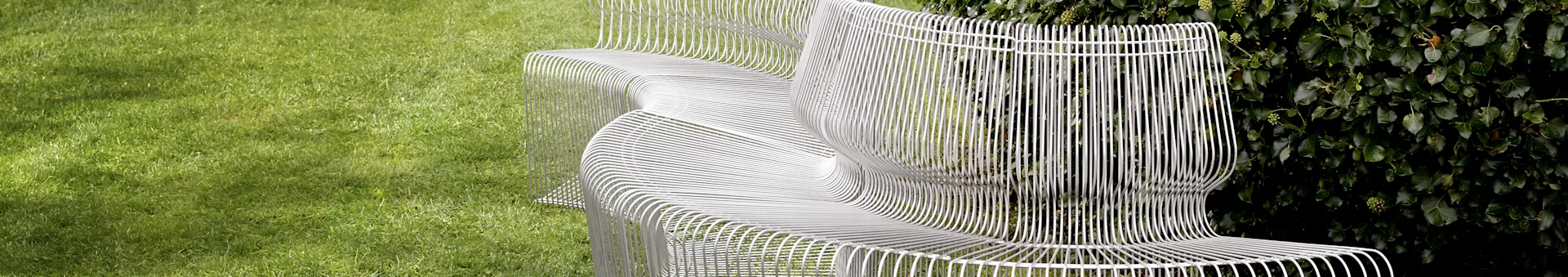 A modern white slatted bench with a wavy design on a green lawn with dark green foliage.