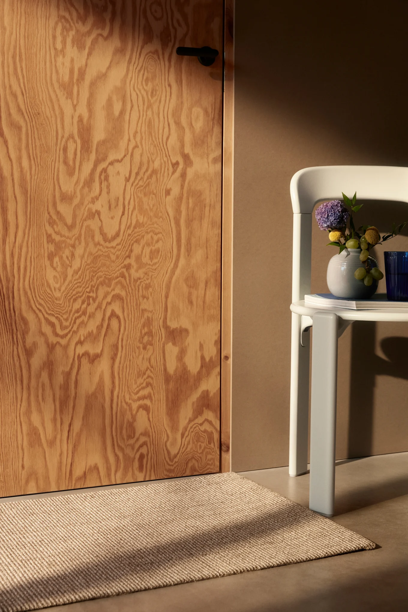 Wooden door and woven rug, with a light chair holding a vase of flowers and grapes.