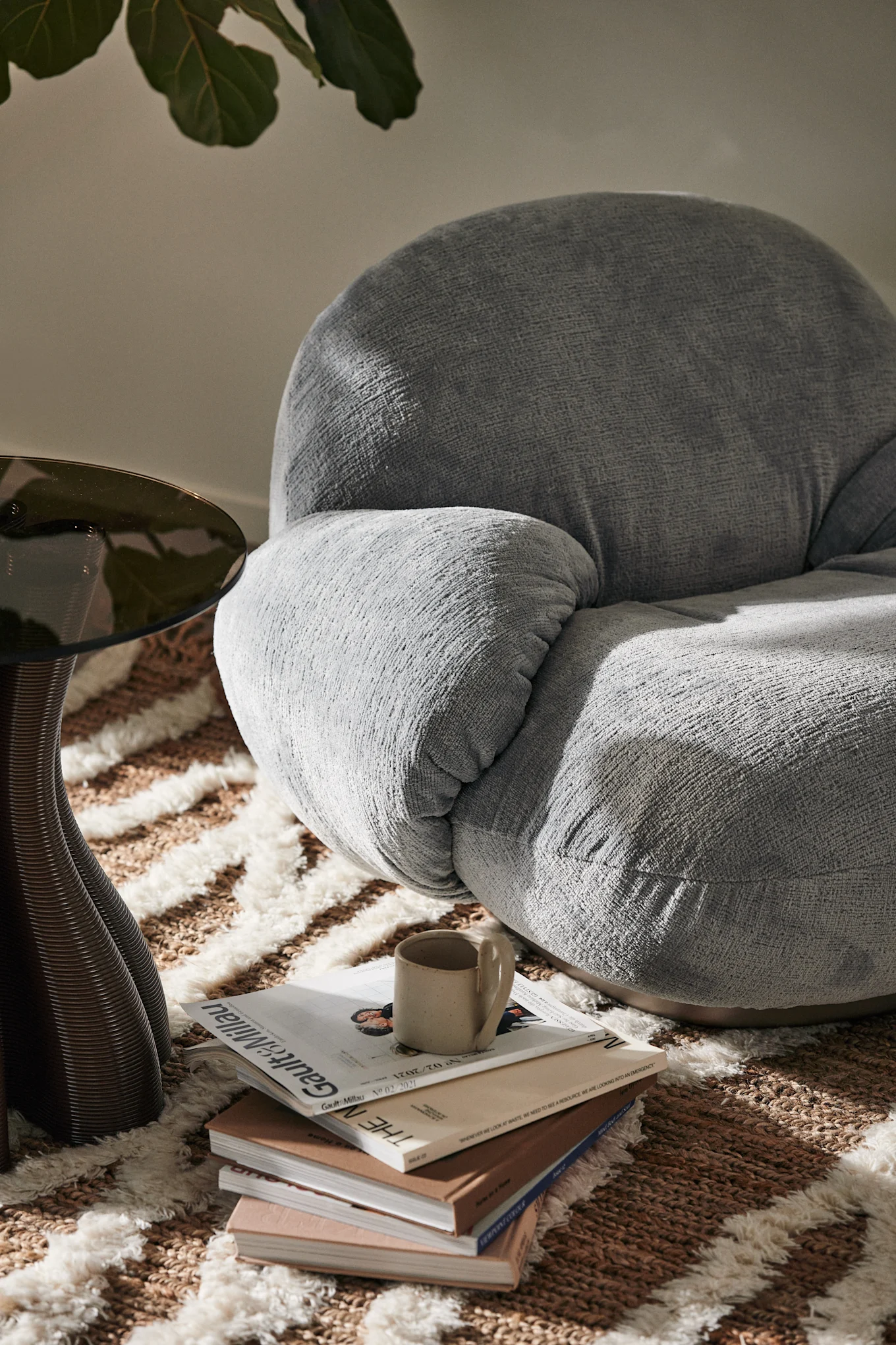 A light grey textured armchair, a dark glass side table, and a stack of books with a ceramic mug on a patterned rug, bathed in sunlight.