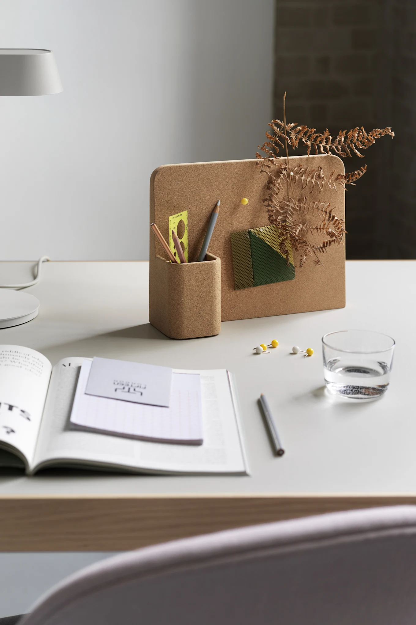 Cork memo board with integrated pen holder holding pencils, a ruler, and a decorative fern. An open book, glass of water, and push pins are on the white desk.