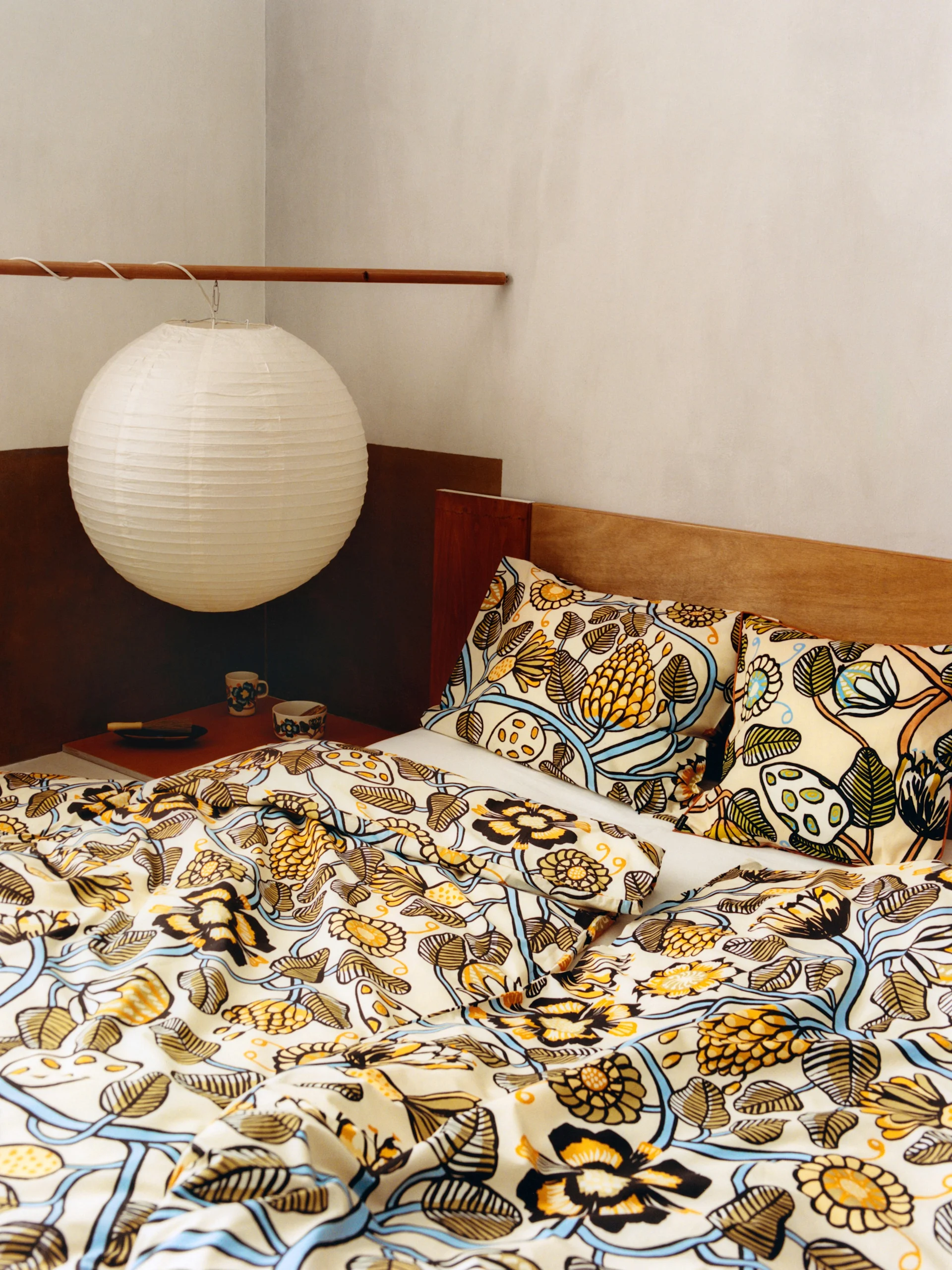 A bed with cream and gold floral patterned bedding and pillows, a wooden headboard, and a white paper lantern.