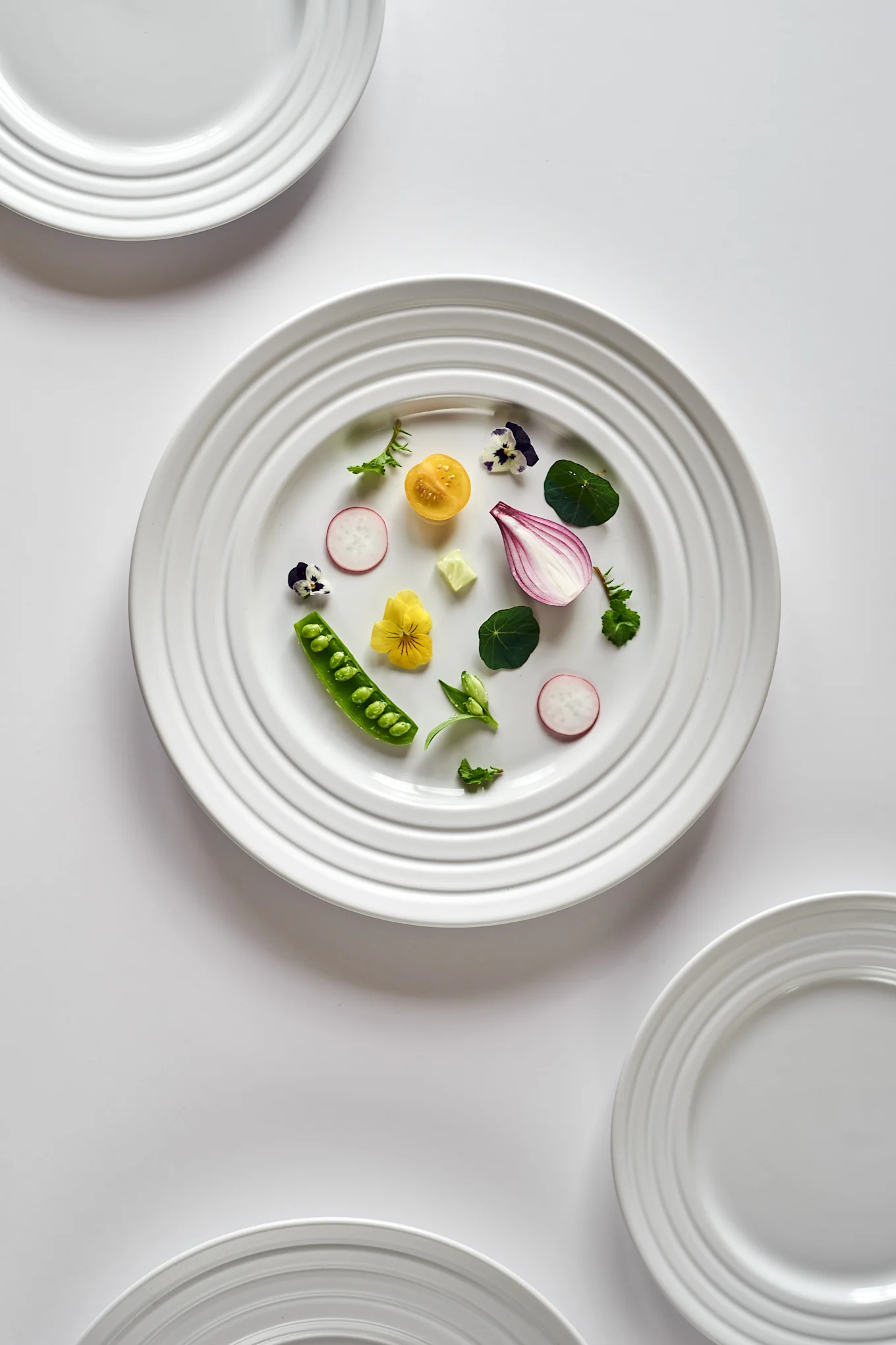 White plate with yellow tomato, red onion, green peas, radish, and edible flowers. Other white plates frame the scene.