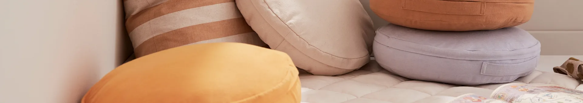Assortment of round and rectangular cushions in mustard, rust, lavender, striped tan, and off-white on a quilted mattress.