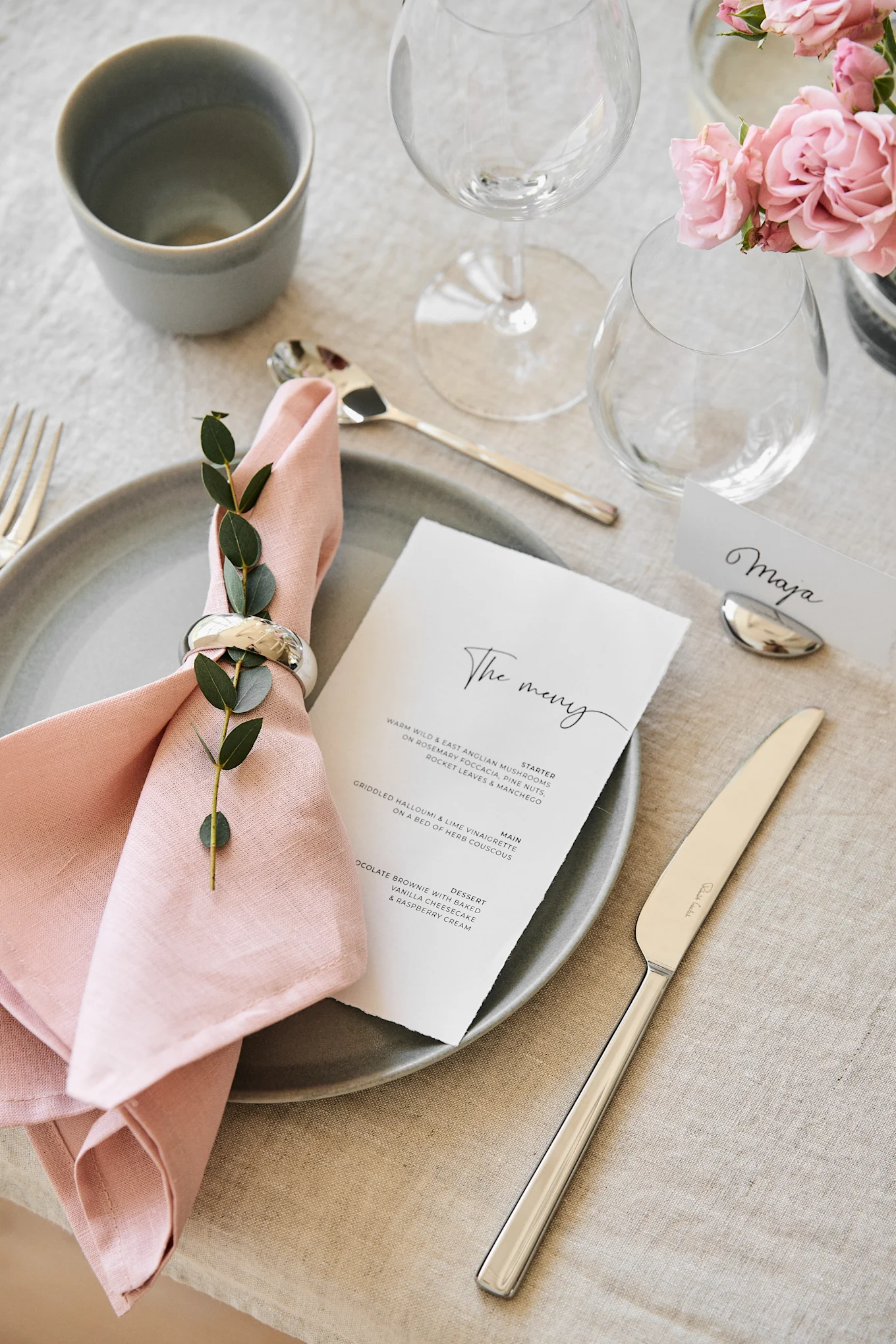 An elegantly set table with a grey plate, pink linen napkin with greenery, a menu, silverware, a 'Maja' name card, and pink roses.