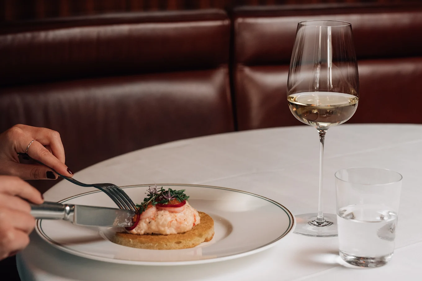 Hands cut a gourmet seafood dish on a blini, garnished with herbs and roe, with wine and water glasses on a white table.