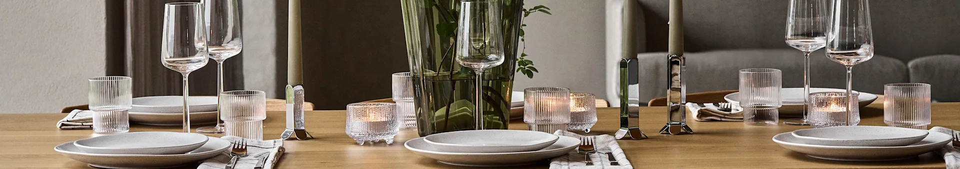 A wooden dining table set with white plates, silver cutlery, clear glasses, lit candles, and a green vase with leaves.