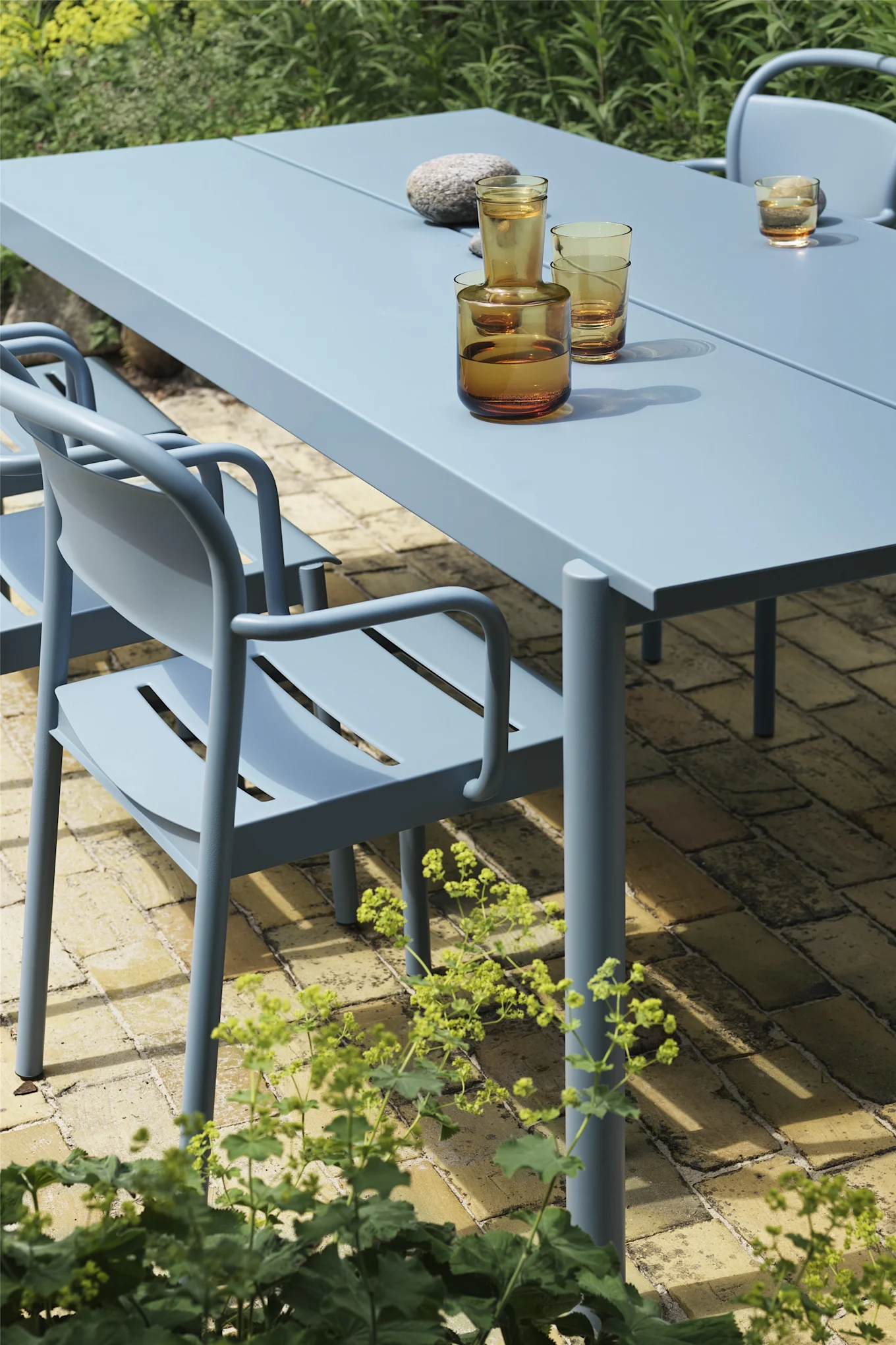 A light blue outdoor dining table with matching chairs on a brick patio, set with amber glasses and a stone.