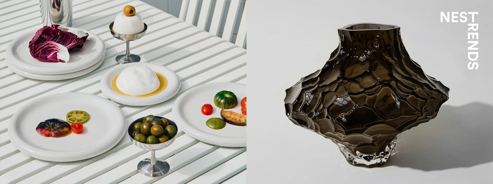 A table with white plates of radicchio, burrata, olives, and colorful tomatoes, next to a textured dark glass vase.