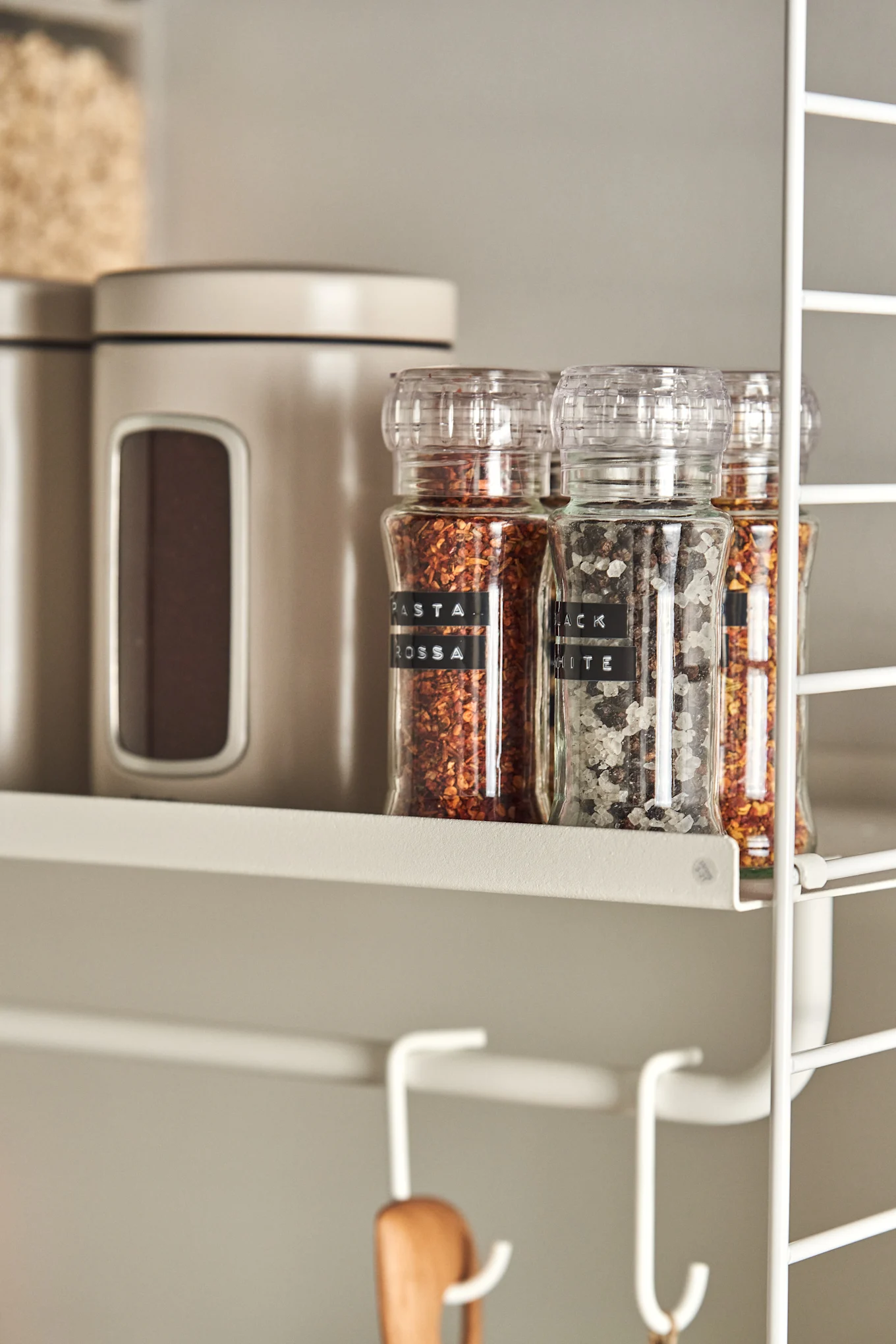 Three spice grinders with chili flakes and salt and pepper sit on a light shelf next to beige containers. White hooks hang below.