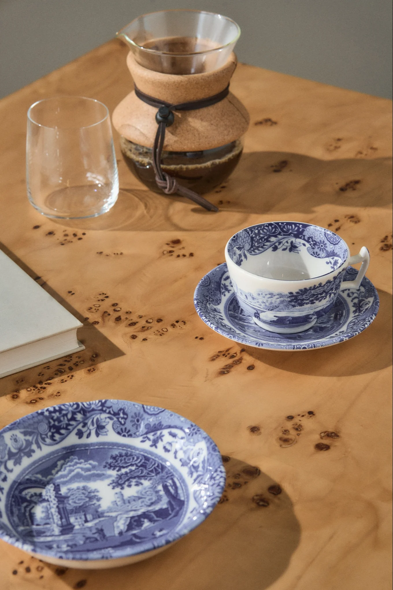 A pour-over coffee maker, an empty glass, a blue and white patterned teacup on a saucer, and a matching plate on a light wooden table.