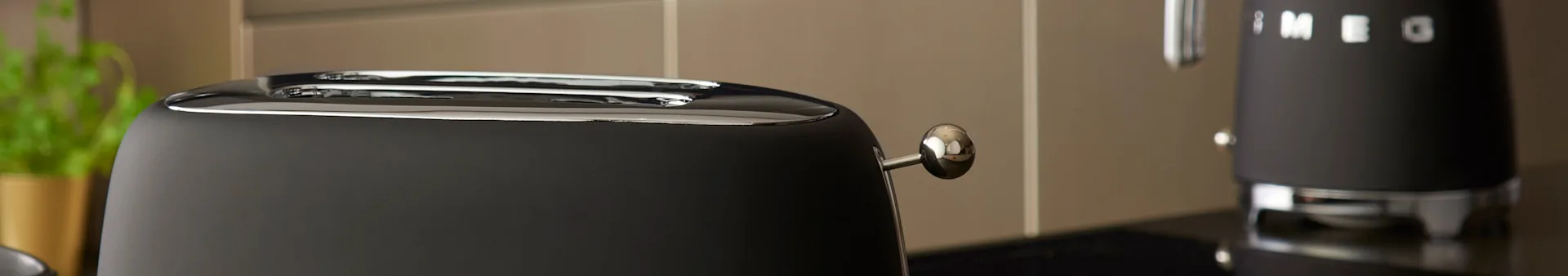 Close-up of a black toaster with two chrome slots and a lever, next to a black kettle in a modern kitchen.