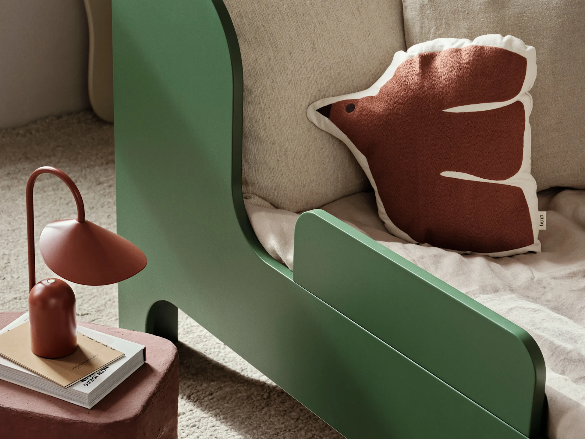 Green child's bed with a rust hedgehog pillow, next to a rust lamp on a stack of books.