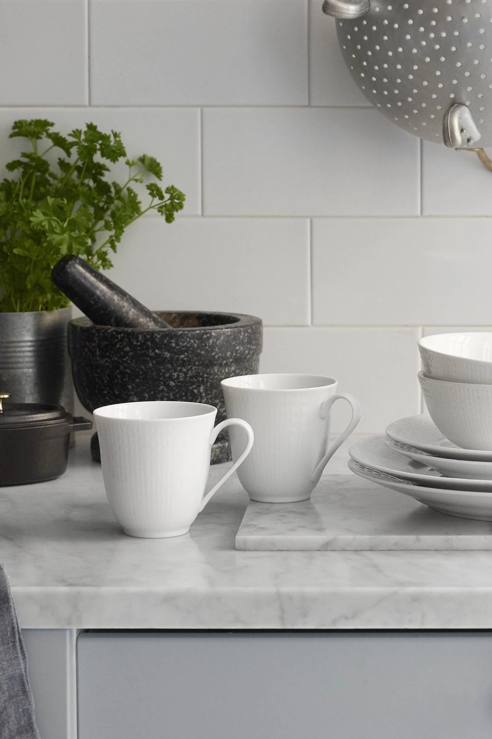 Two white ribbed mugs and stacked bowls and plates on a marble kitchen counter with a mortar and parsley.