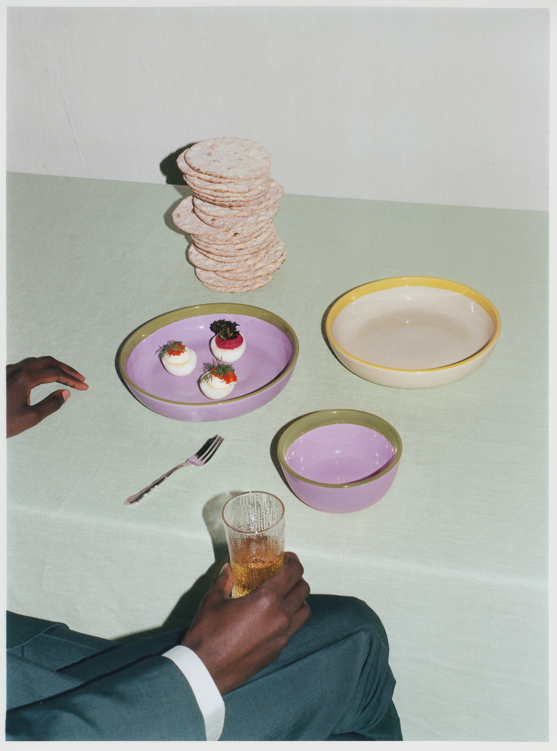 Deviled eggs and flatbread on a table with purple and beige ceramic bowls. A hand holds a textured glass.