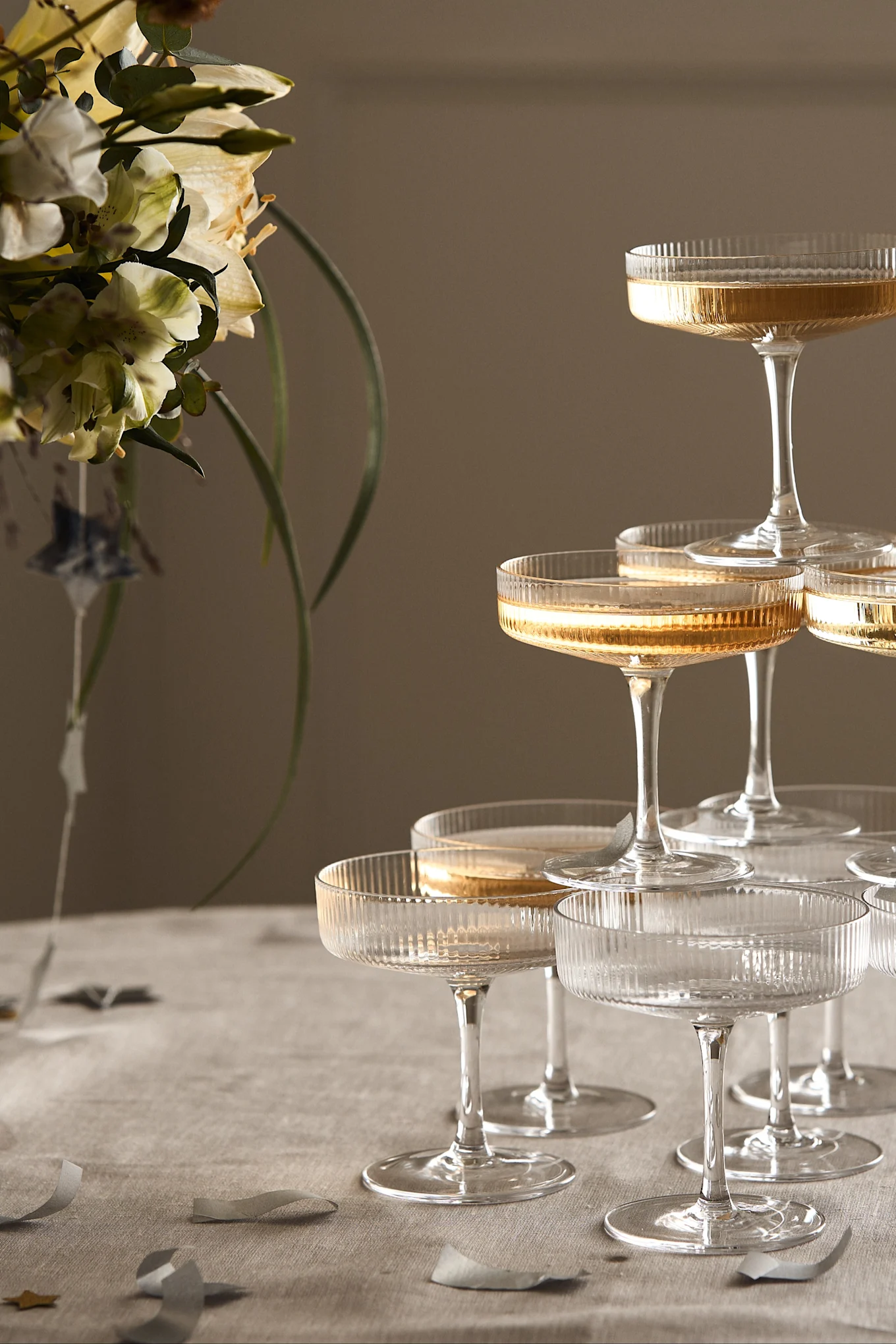 A champagne tower of ribbed coupe glasses filled with golden liquid, with light flowers and confetti on a tablecloth.