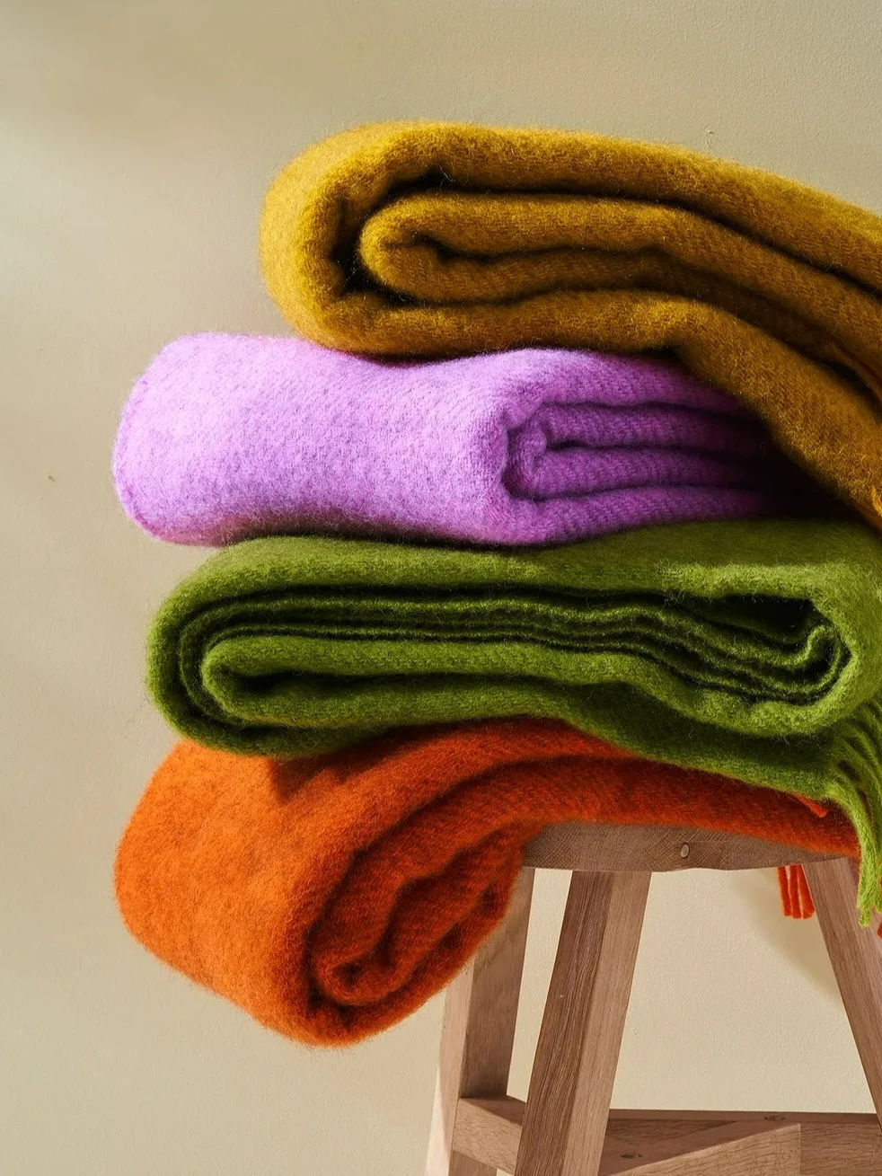 A stack of four soft, textured blankets in mustard yellow, lilac, olive green, and burnt orange on a wooden stool.
