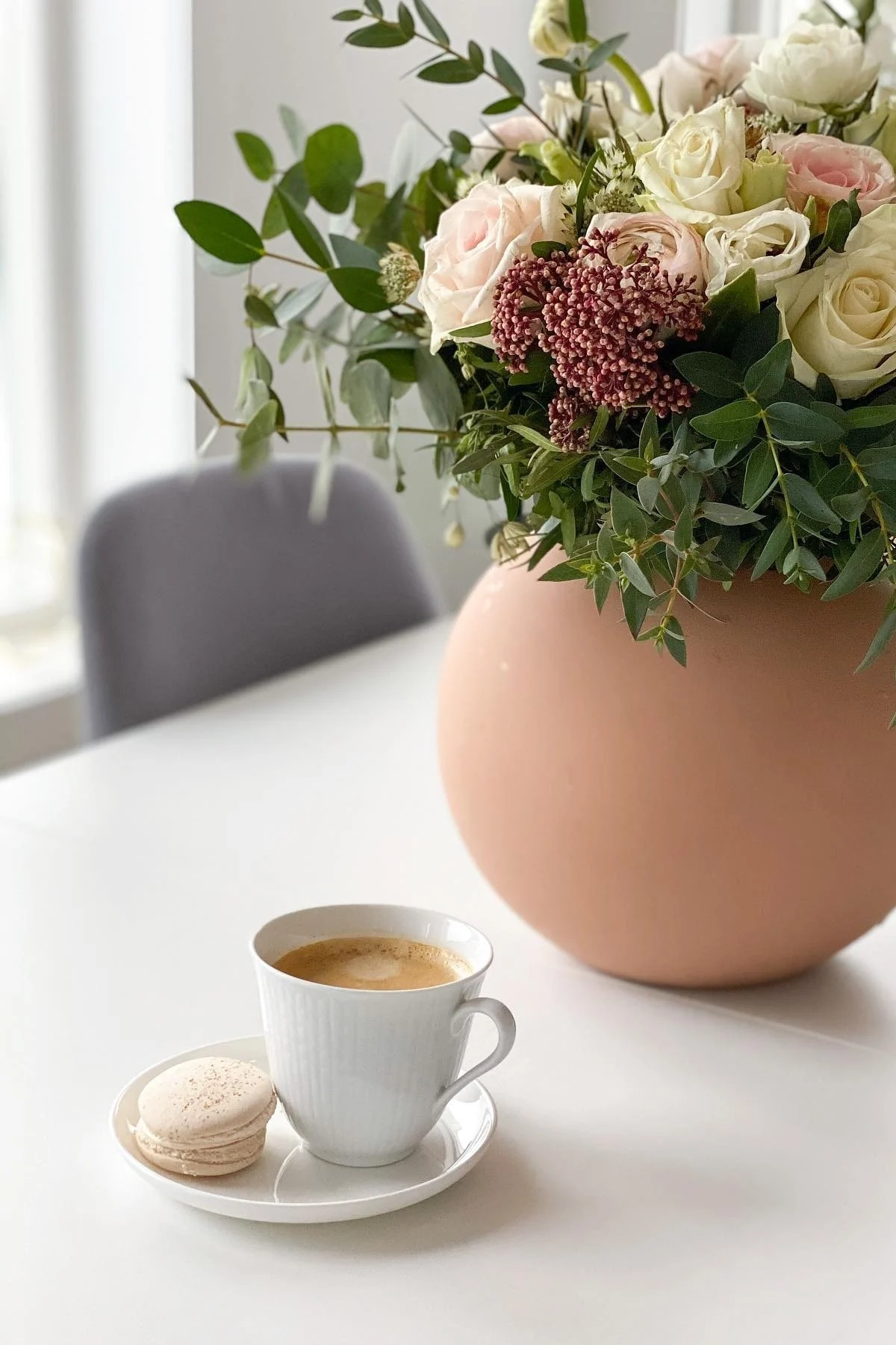 A bouquet of pink and white roses with greenery in a peach vase, next to a coffee cup and macaroon on a white table.