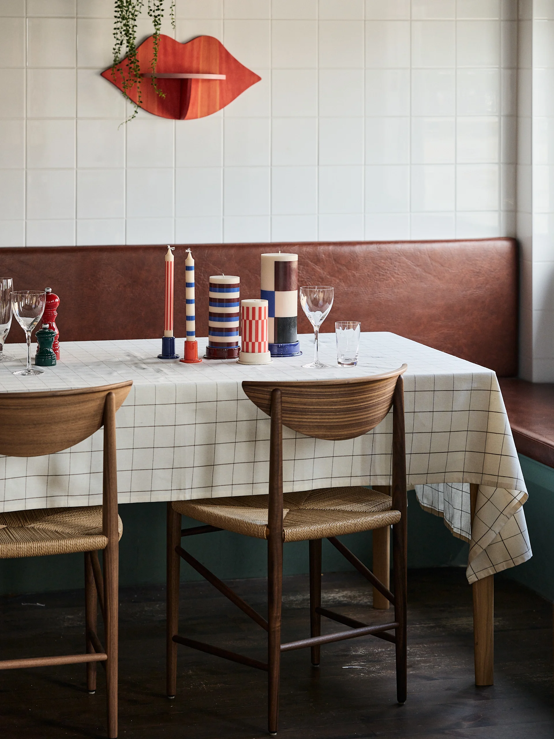 A dining table set with candles in geometric patterns, two wooden chairs, and a grid tablecloth, with a red lip-shaped wall decor in the background.