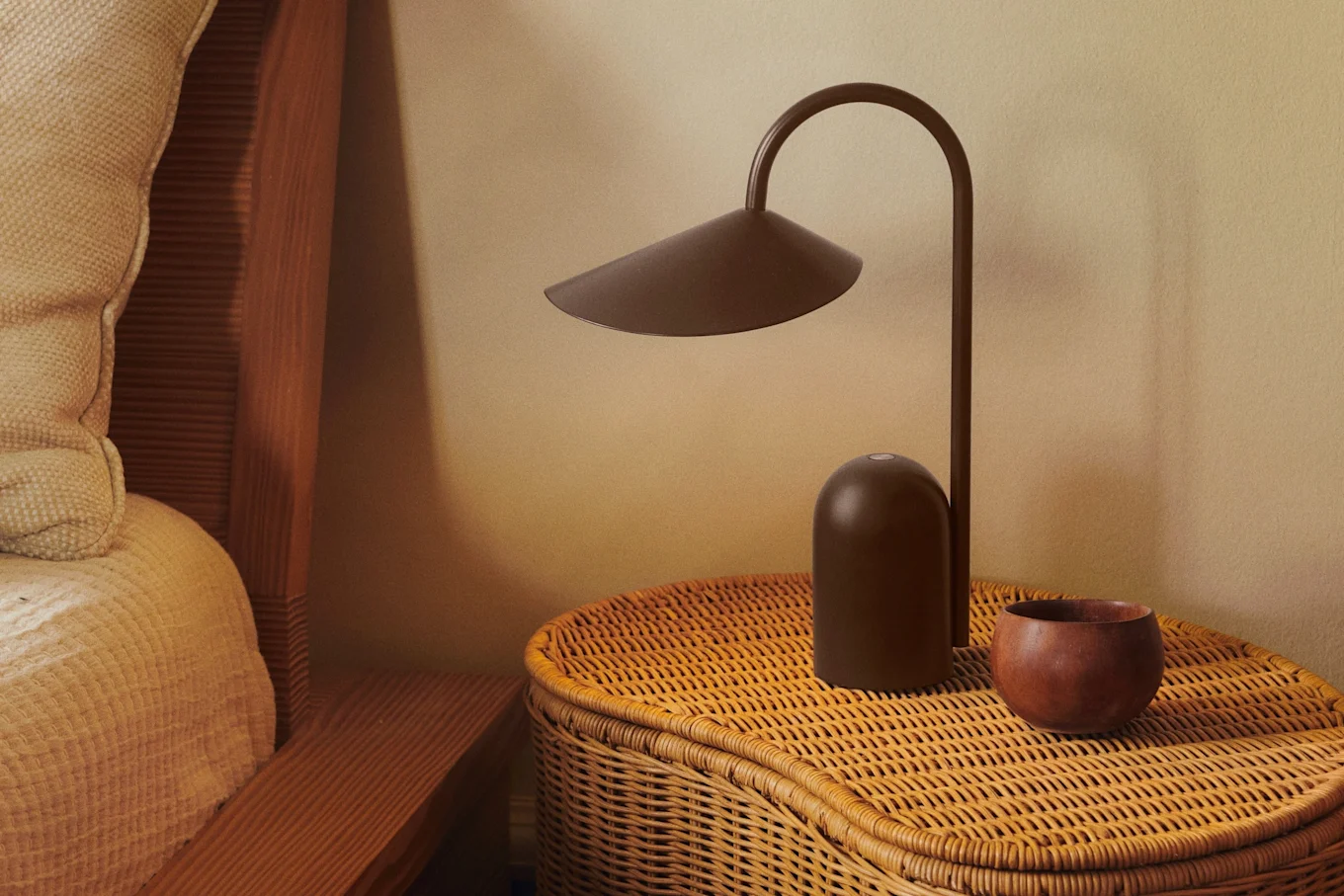 A dark brown modern lamp and a small wooden bowl sit on a woven wicker bedside table next to a bed with a textured beige blanket.