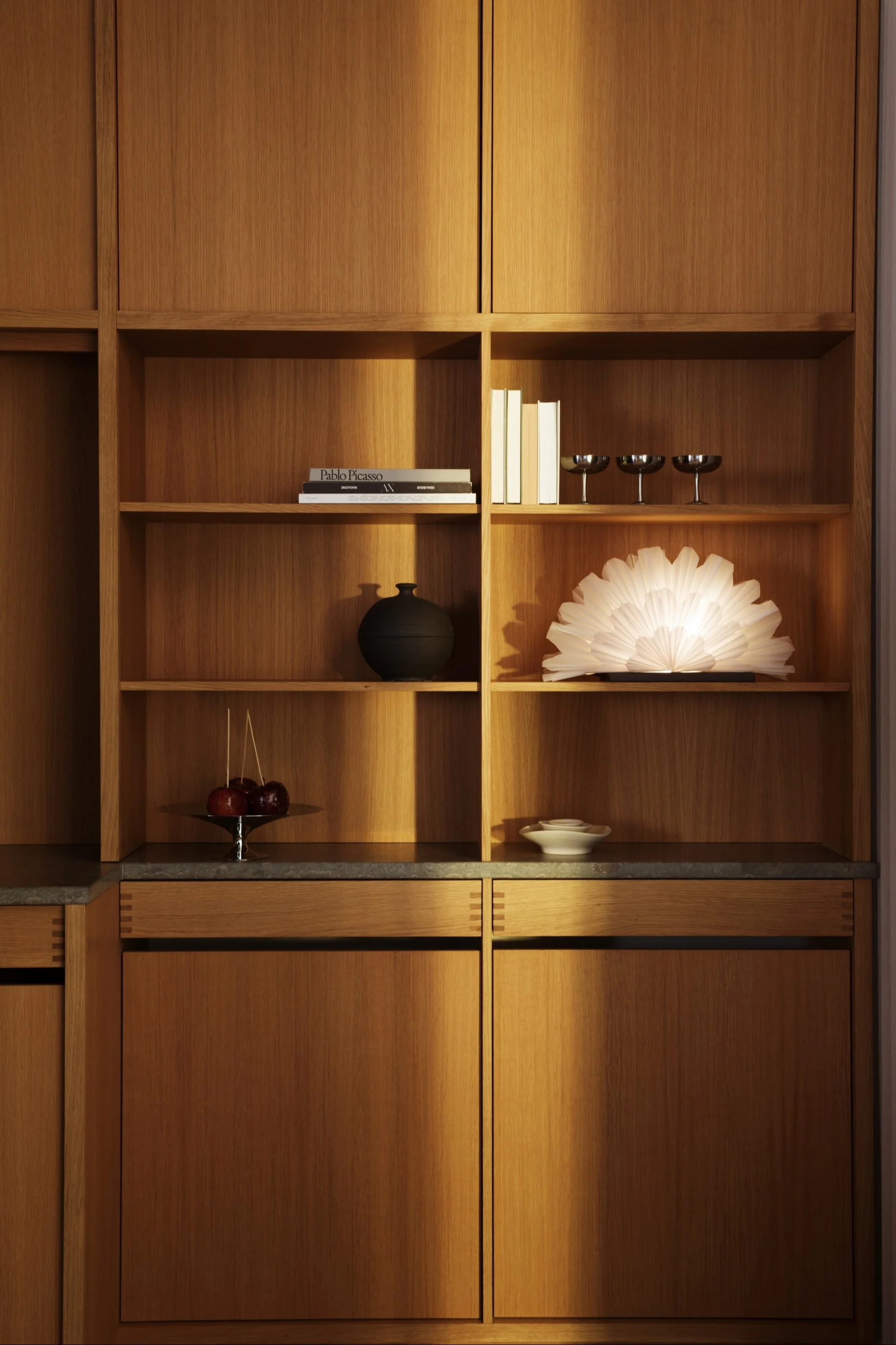 A modern built-in wooden cabinet with open shelves displaying books, a black vase, a glowing fan-like lamp, candy apples, and silver coupes.