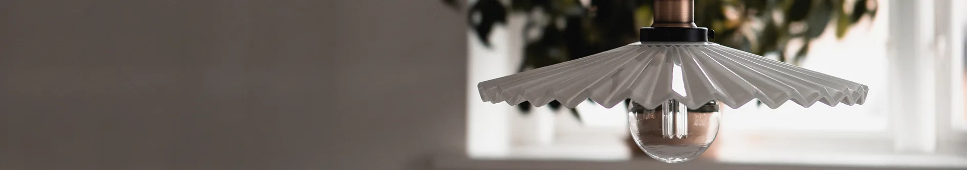 A modern pendant light with a pleated white shade and a clear glass bulb, against a blurred indoor background.